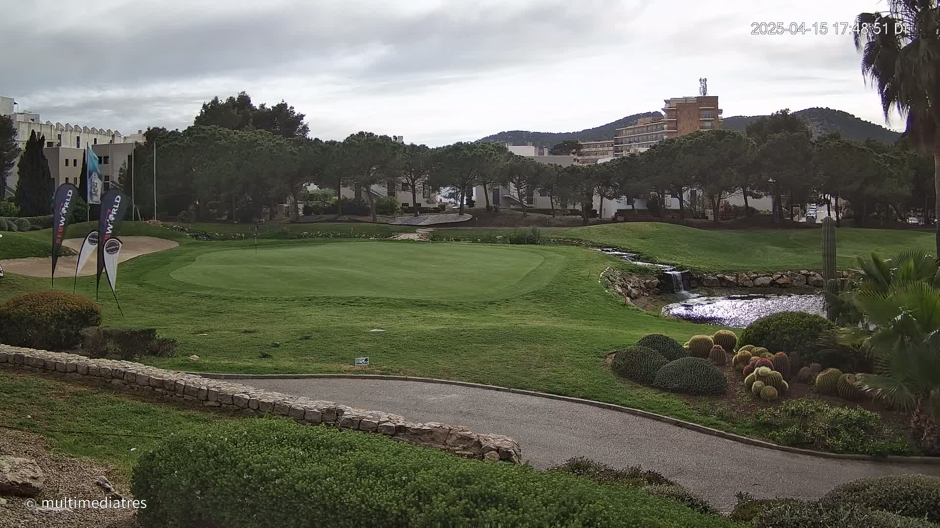 A golf course green with a small pond and waterfall is situated near buildings and trees under an overcast sky.