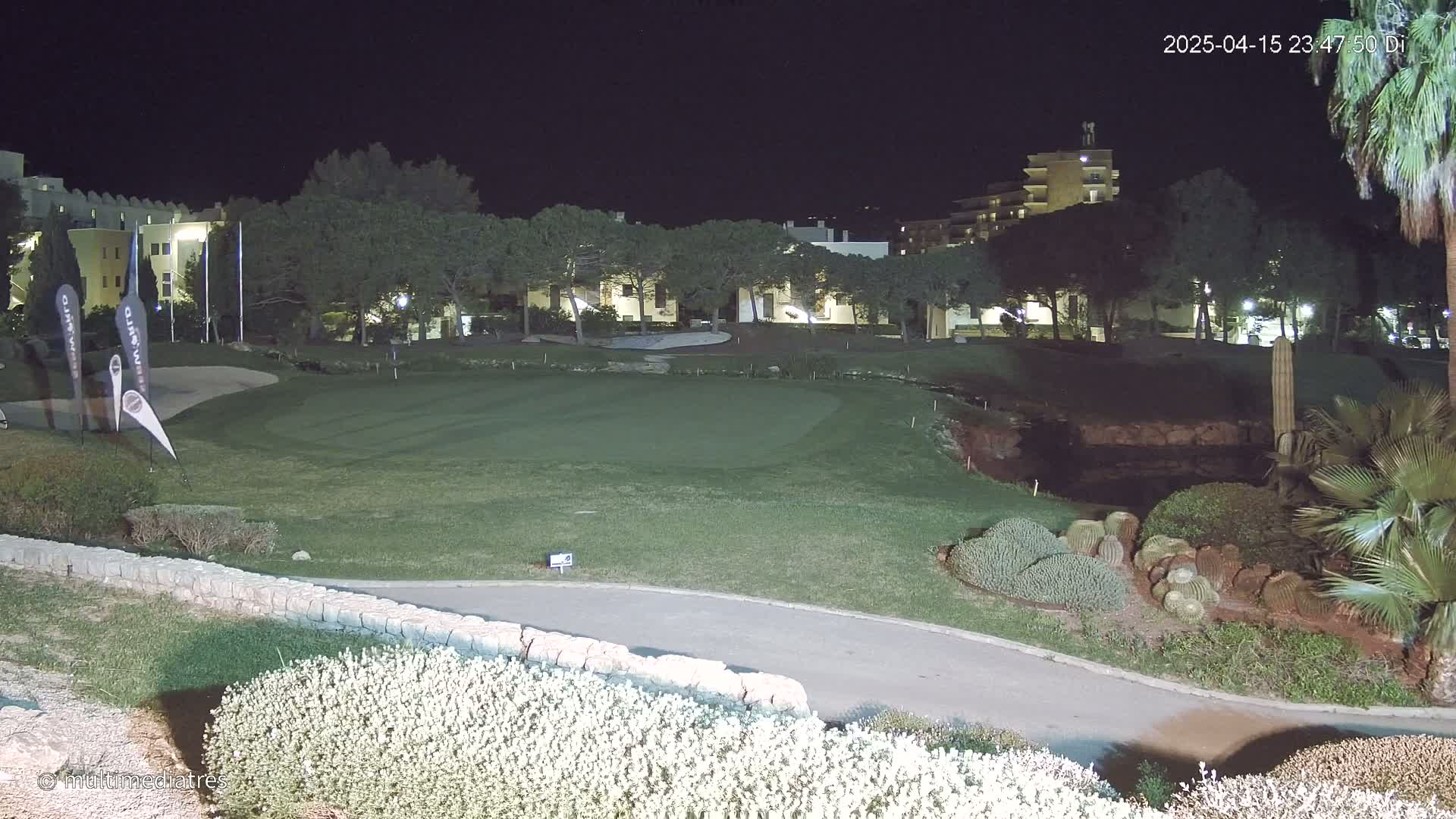 A nighttime view of a golf course green, bordered by low stone walls and landscaping, with buildings and trees illuminated in the background.