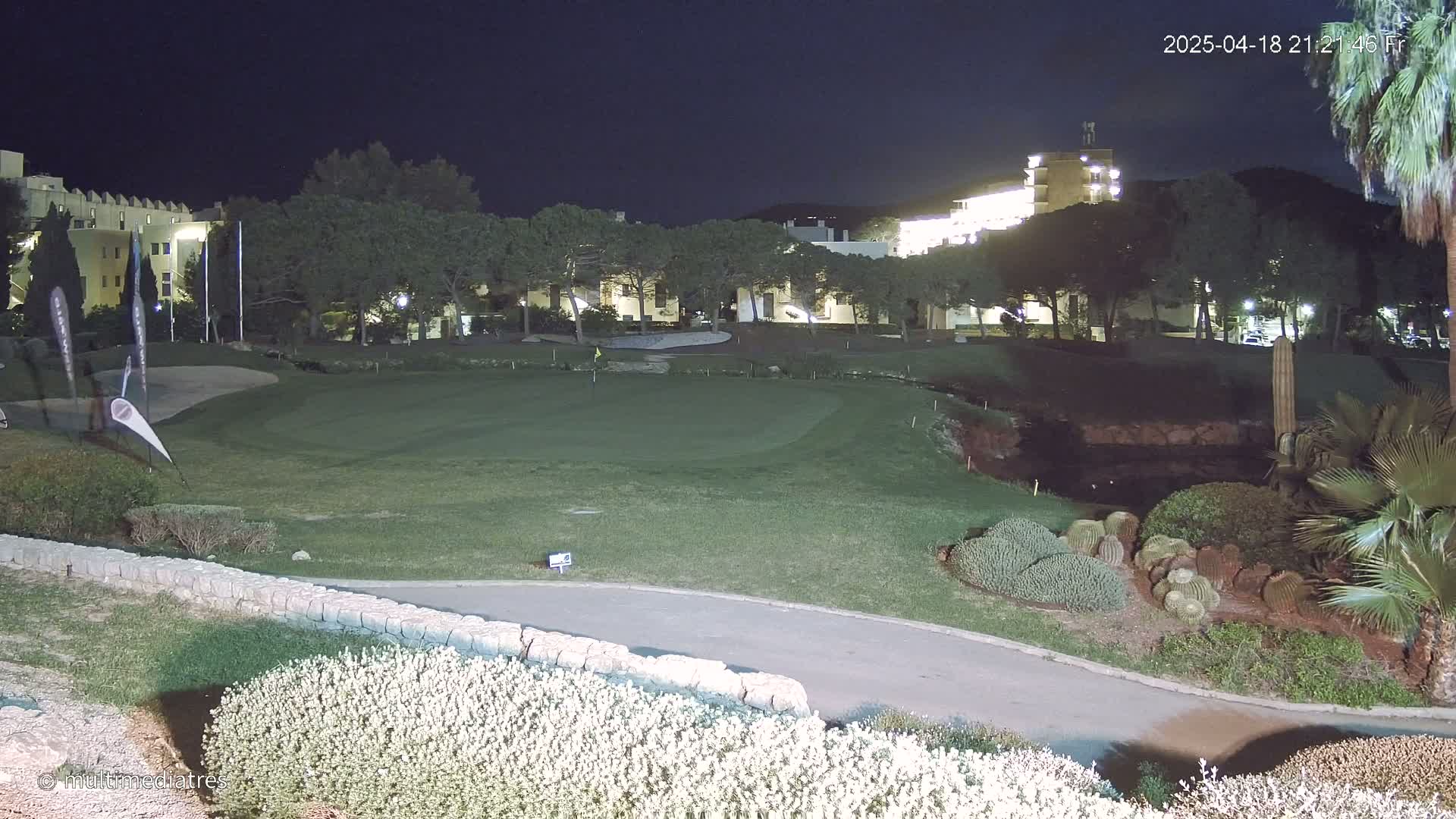 A nighttime view of a golf course green, bordered by shrubs and trees, with a lit building in the background under a dark sky.