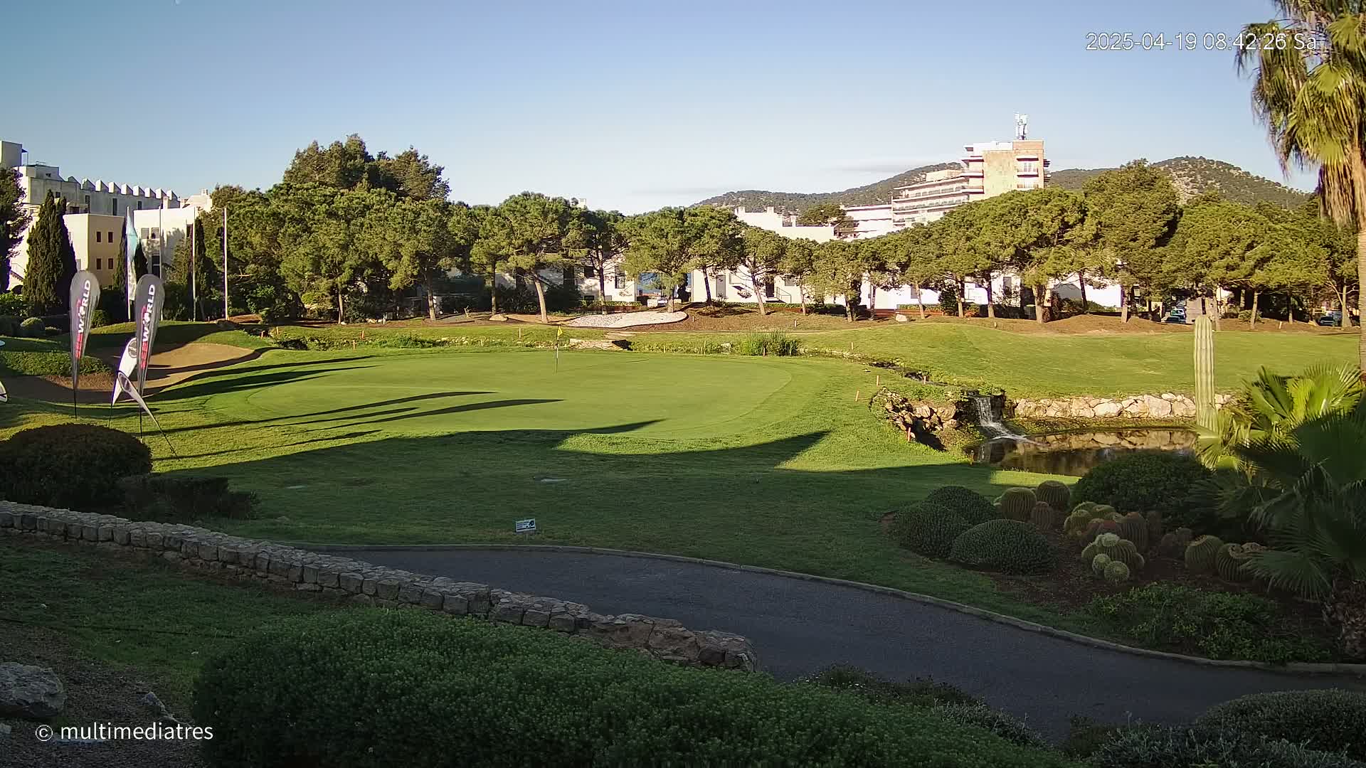 A sunny day reveals a golf course with a green, a small pond, and several buildings in the background, surrounded by lush greenery.