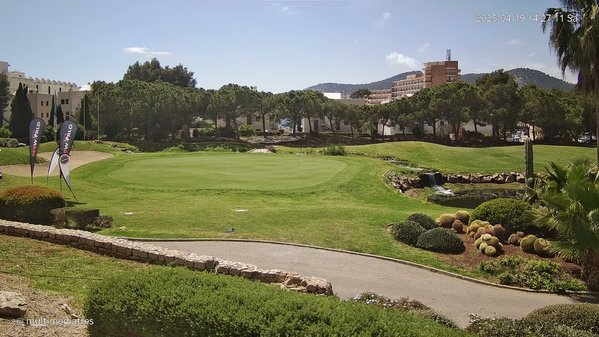 A golf course green with a small pond and a stone wall, under a sunny sky, is surrounded by lush greenery and buildings in the background.