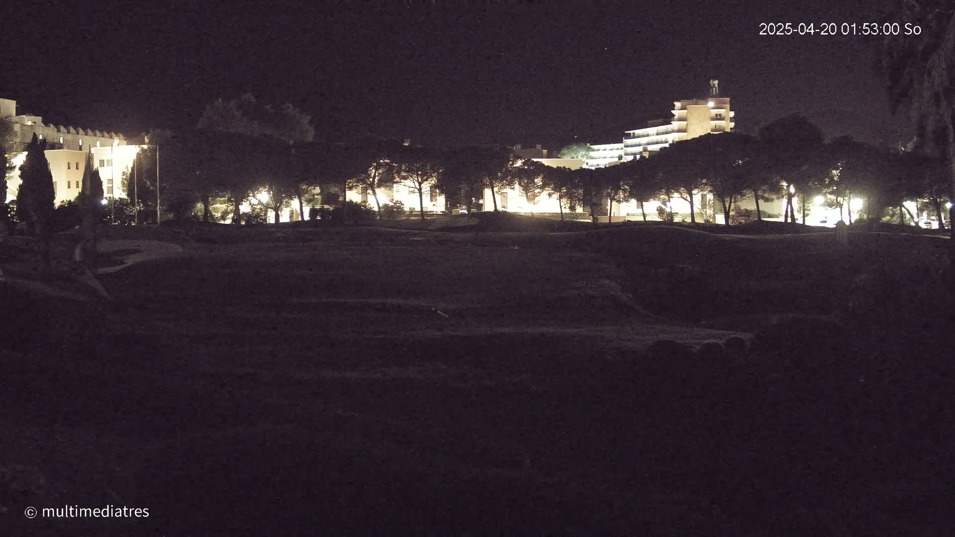 A dark, nighttime scene shows a line of illuminated buildings beyond a row of trees and a large, mostly dark, field in the foreground.