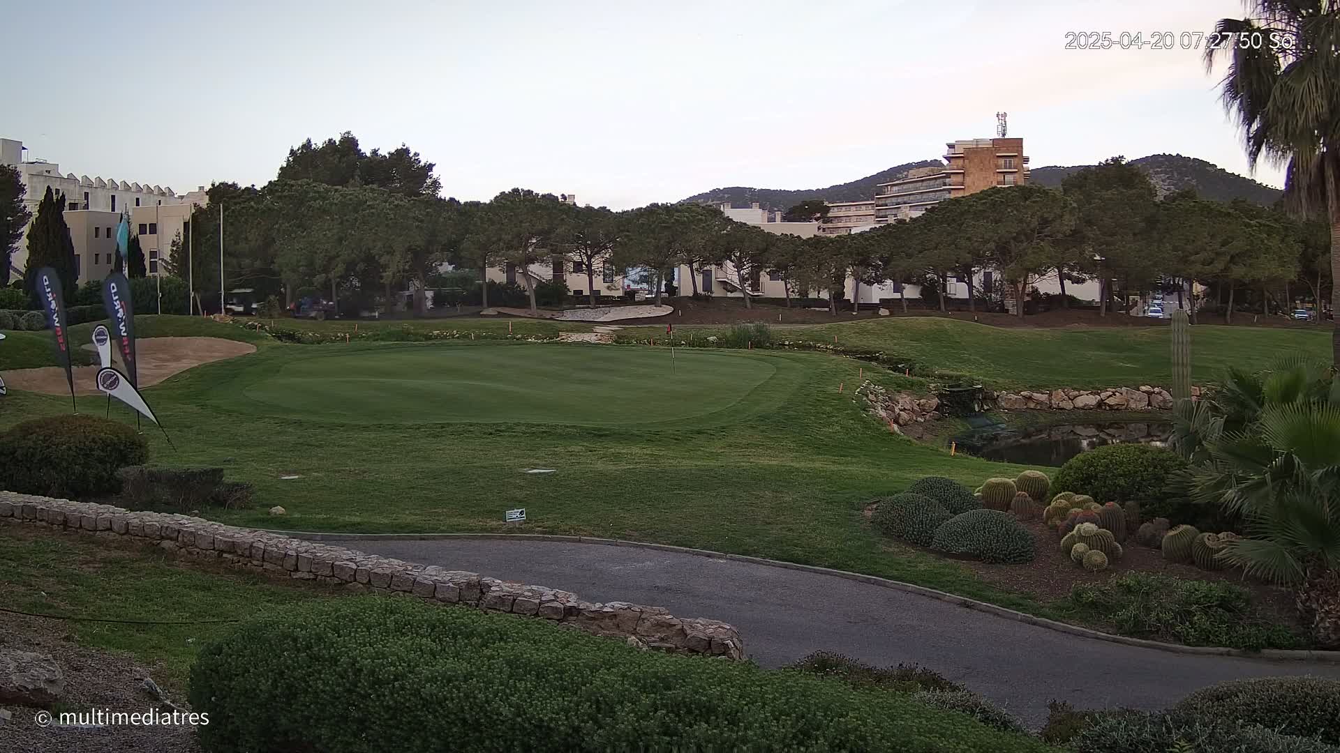 A golf course green, bordered by a low stone wall and various shrubs and cacti, is situated near residential buildings under a twilight sky.