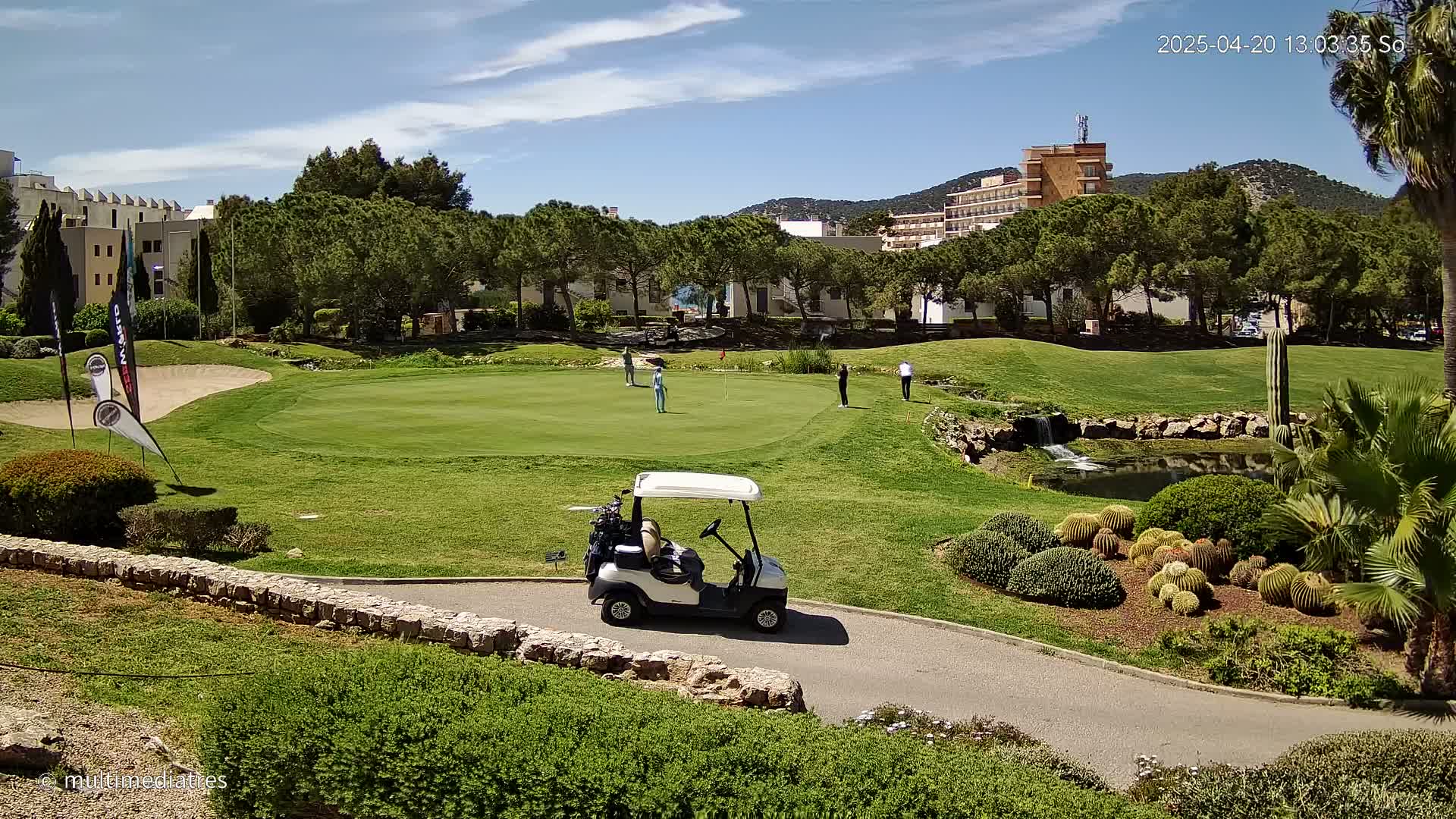 A golf course with several people playing, a golf cart on the path, and a small pond with landscaping featuring cacti in the sunny weather.