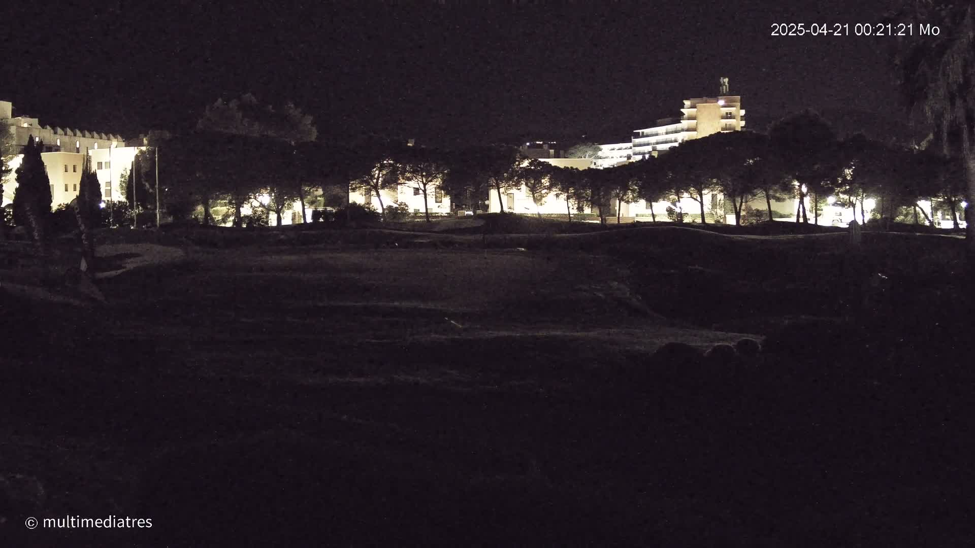 At night, illuminated buildings are visible behind a line of dark trees, in front of a darker, empty field.