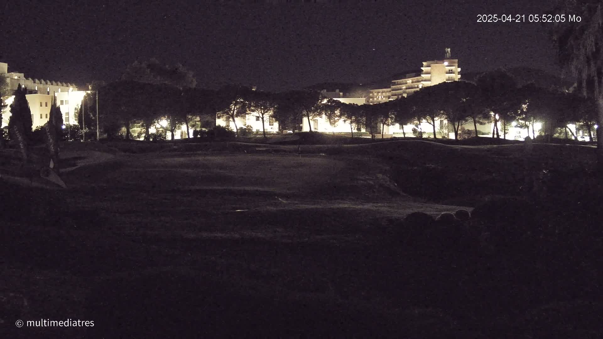 A dark, nighttime view of a golf course with illuminated buildings and trees in the background.