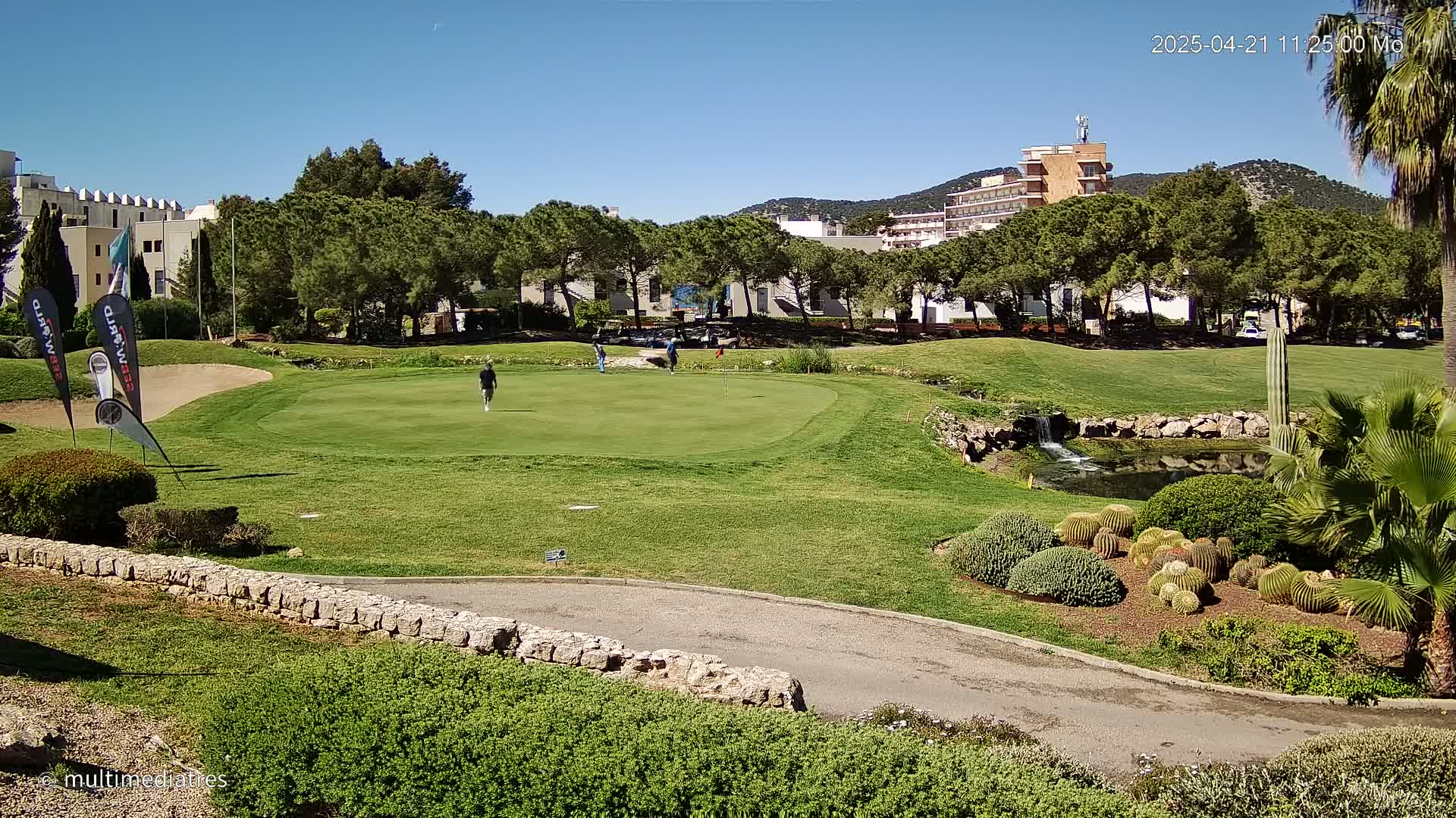 A golf course with several golfers on a green, a small pond, and landscaping featuring cacti and other desert plants, under a clear blue sky.