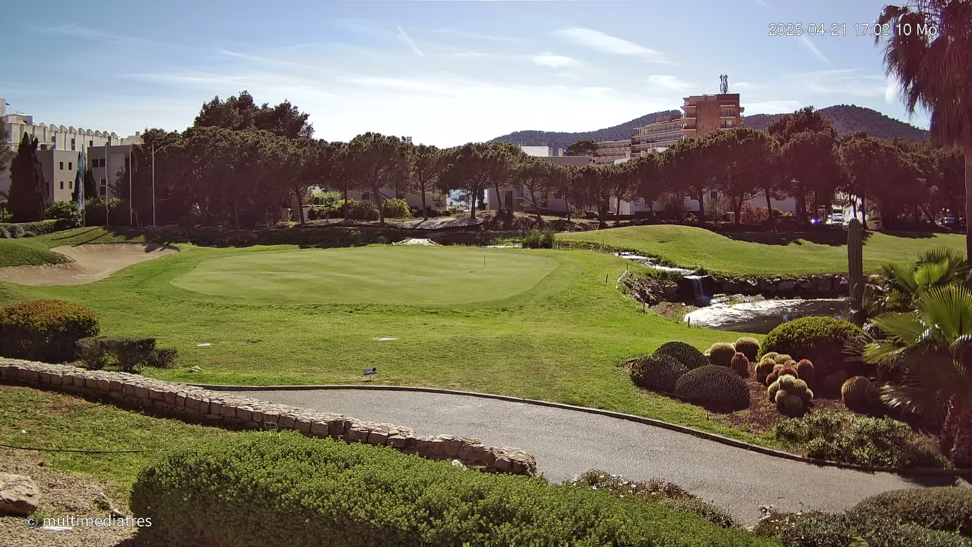A sunny golf course with a green, sand trap, small pond, and landscaping featuring cacti and shrubs, is situated near buildings and a mountain in the background.