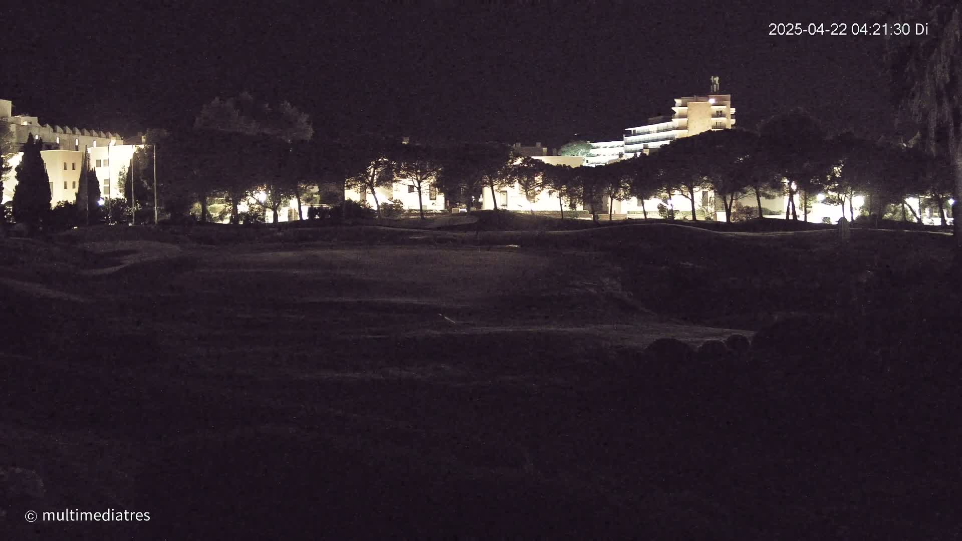 At night, illuminated buildings are visible beyond a dark, uneven expanse of land, possibly a field or golf course, lined with trees.