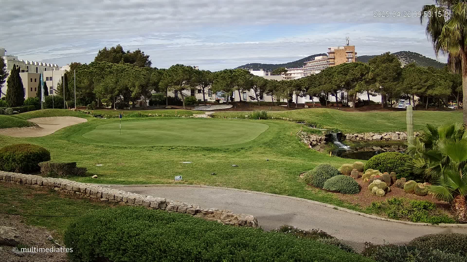 A golf course green with a sand trap, a small waterfall, and desert landscaping is situated near buildings under a cloudy sky.