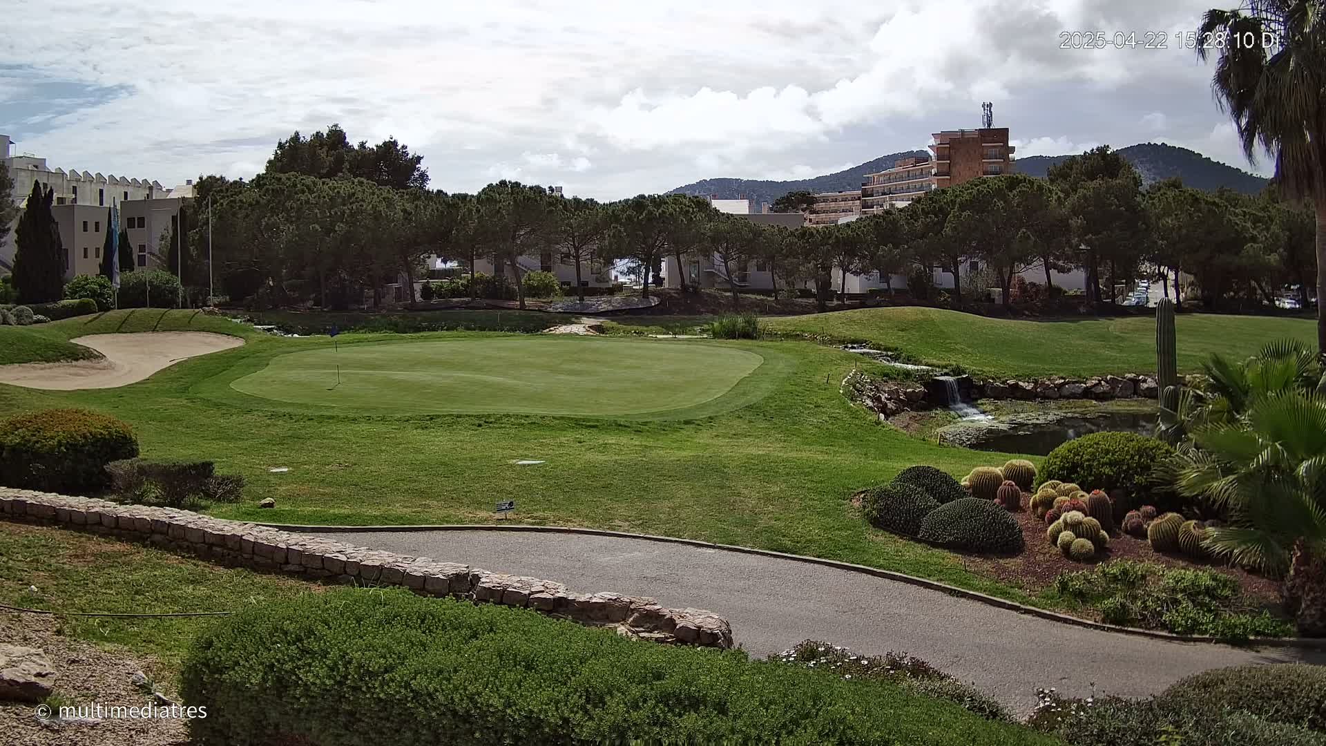 A golf course green with a sand trap and a small waterfall feature is visible under partly cloudy skies, with buildings and trees in the background.