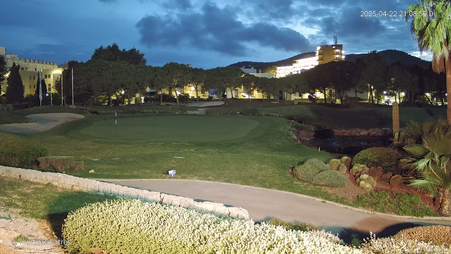 A well-lit golf course green at twilight, with low-lying landscaping and buildings in the background under a partly cloudy sky.