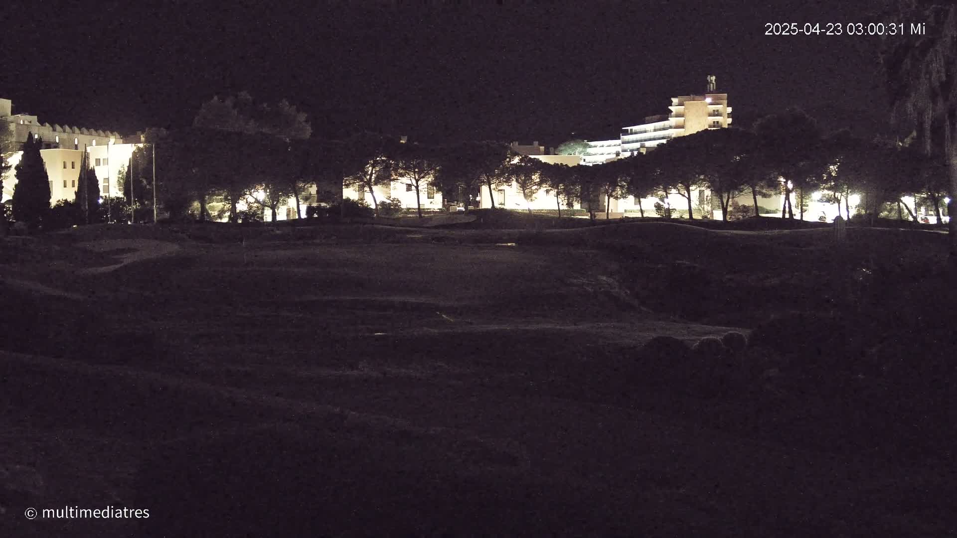 A dark, nighttime scene shows a row of illuminated buildings behind a line of trees and a large, dark, uneven expanse of land in the foreground.
