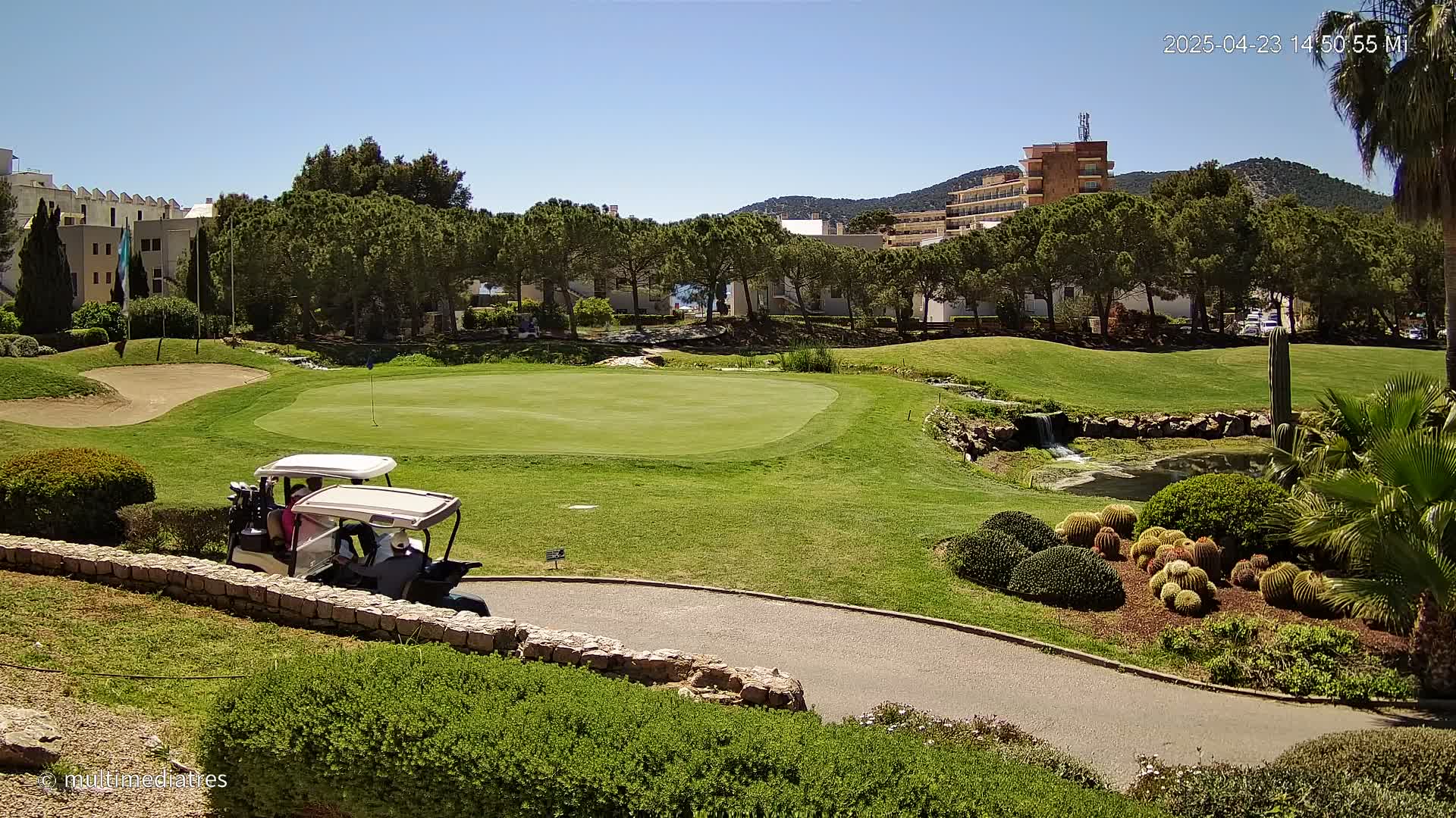 A golf course with a golf cart and putting green, under a sunny sky, is surrounded by landscaping, buildings, and trees.
