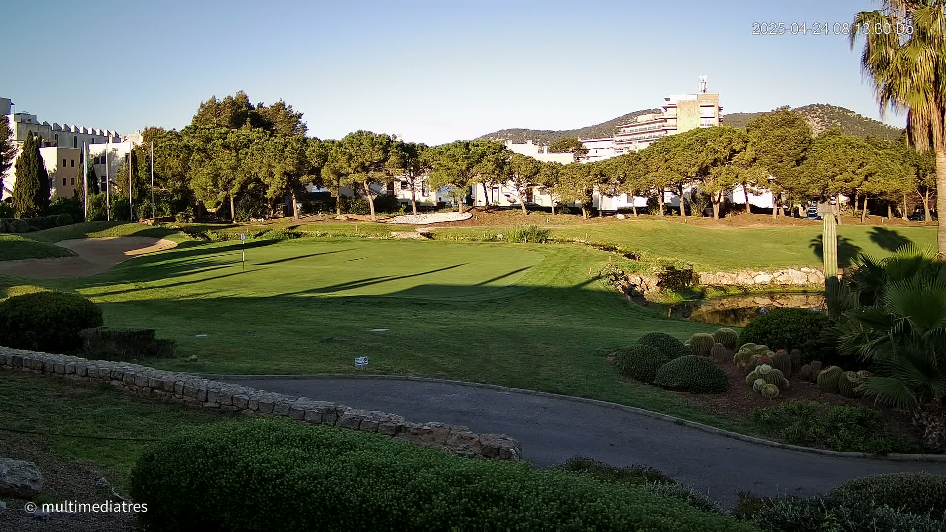 A golf course green with sand traps and a small pond is situated near buildings and a row of trees under a clear, sunny sky.