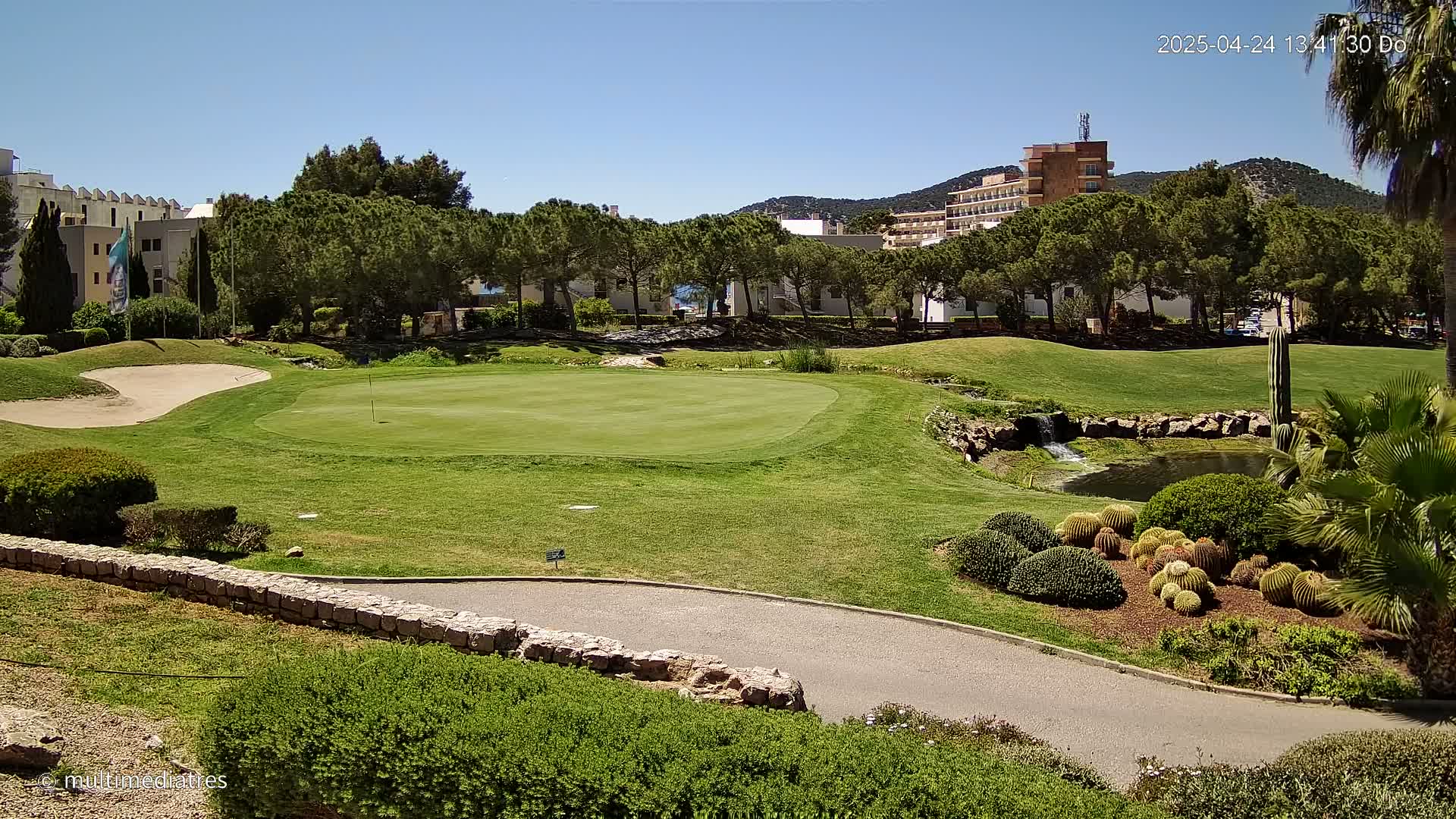 A golf course putting green with a sand trap and a small pond, surrounded by lush greenery, buildings, and palm trees under a clear blue sky.