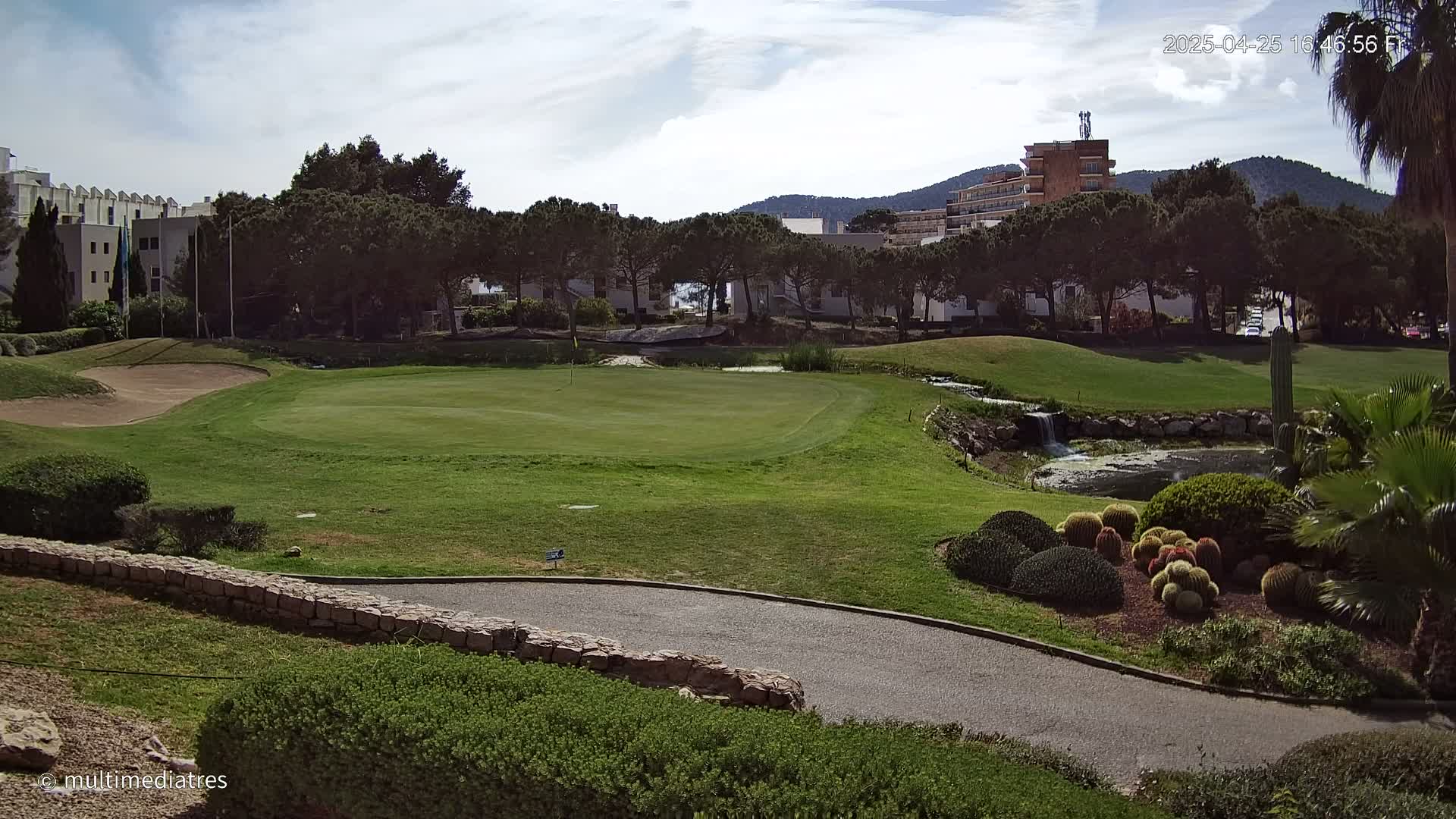 A green golf course with a sand trap, a small pond with a waterfall, and a line of trees behind it under a mostly sunny sky.