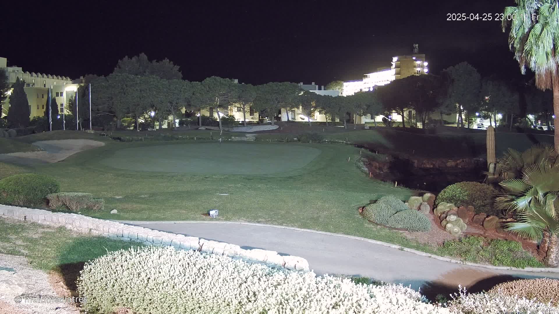 A nighttime view of a golf course putting green, bordered by landscaping and illuminated buildings in the background.