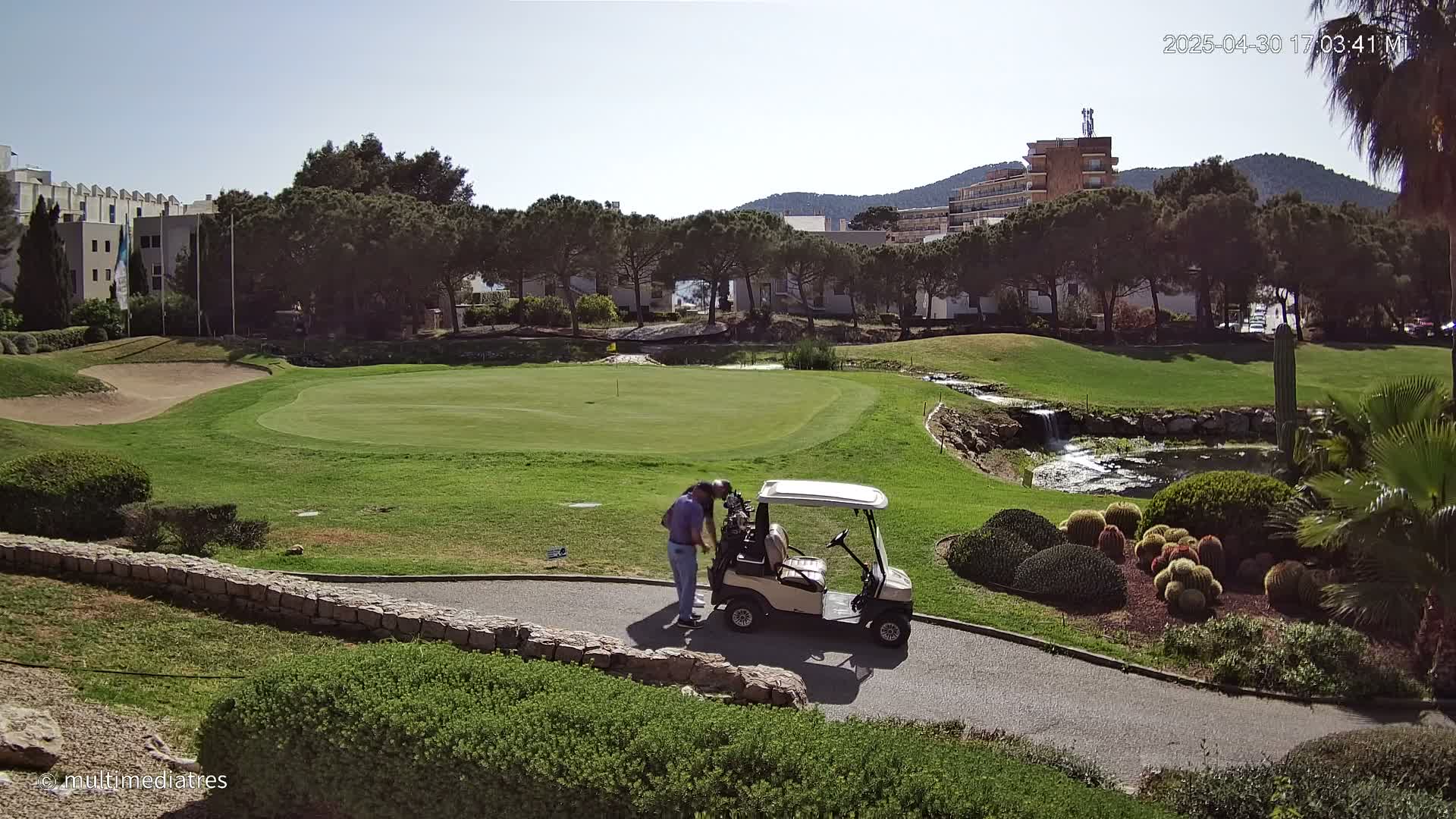 A golfer examines a golf cart on a sunny golf course with a small pond and landscaping features.