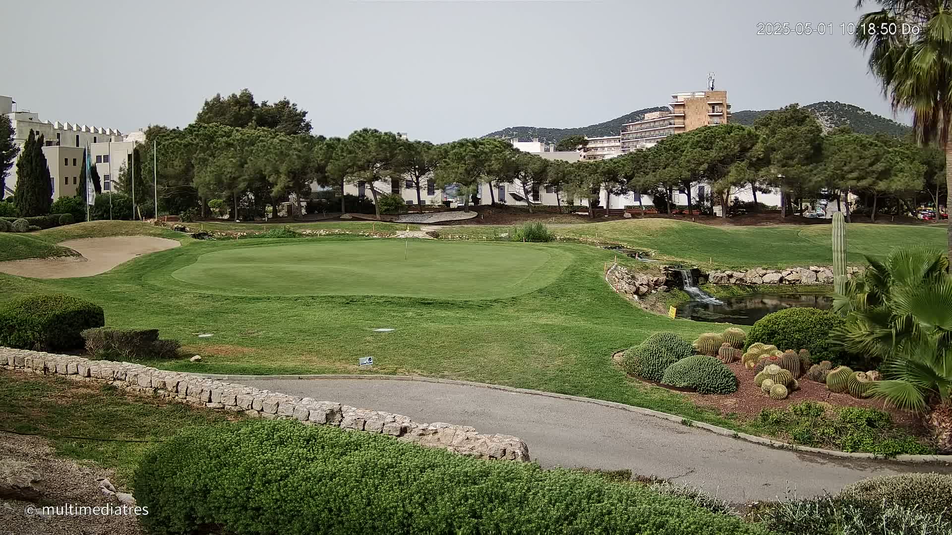 A golf course putting green with a small pond and landscaping, situated near buildings and under a cloudy sky.