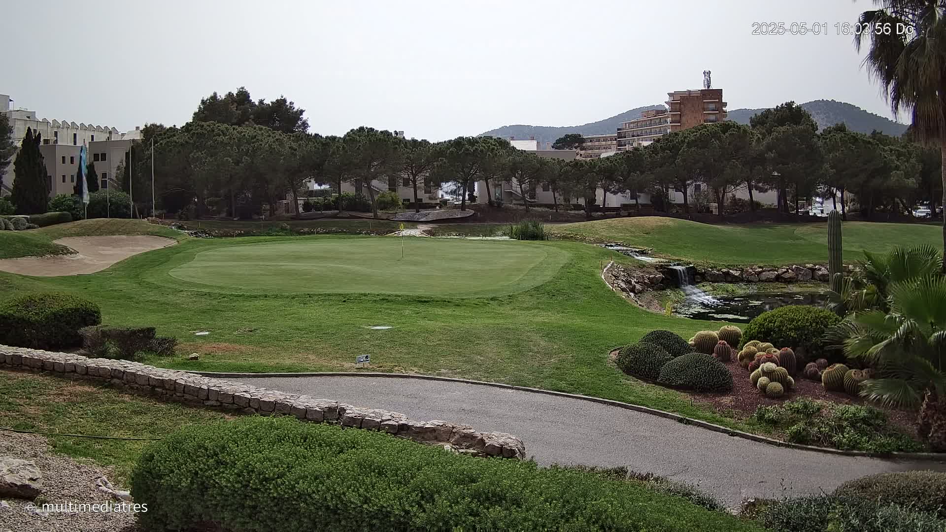 A golf course putting green with a small waterfall and a collection of cacti is visible under an overcast sky.