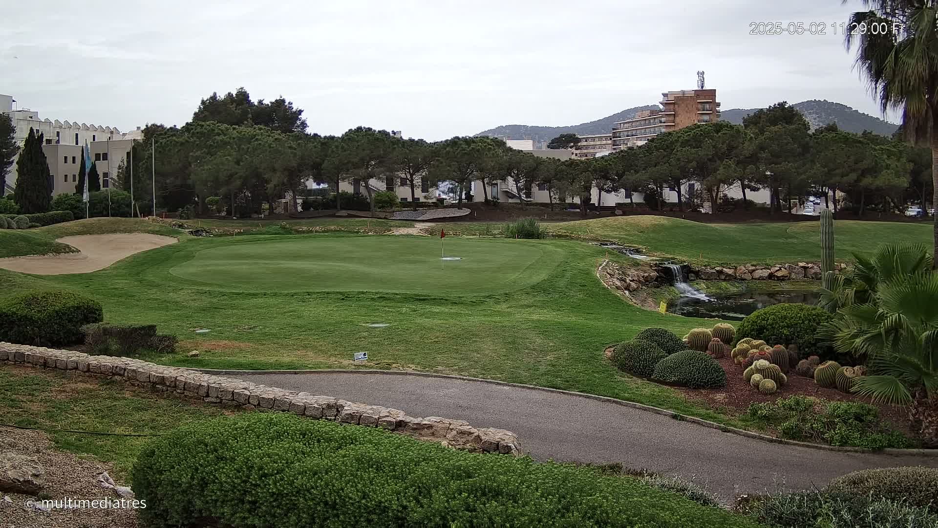 A golf course putting green with a small waterfall and pond, surrounded by lush landscaping and buildings in the background under an overcast sky.
