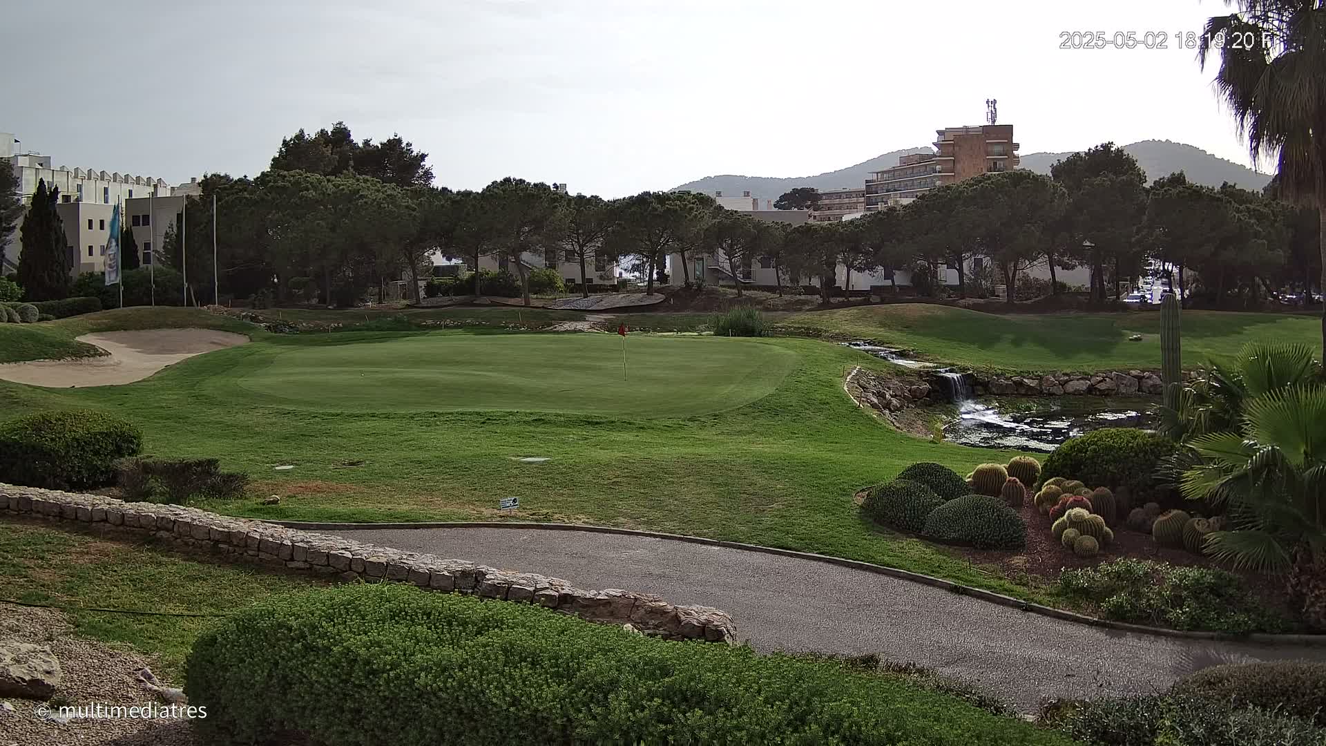 A golf course green with a sand trap, a small waterfall, and a variety of lush landscaping under a cloudy sky.