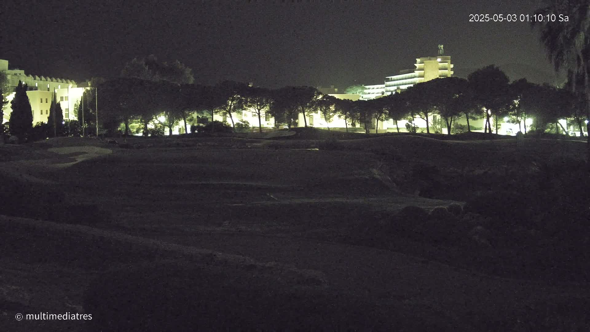 At night, a line of illuminated trees separates a dark, uneven foreground from a dimly lit building in the background.