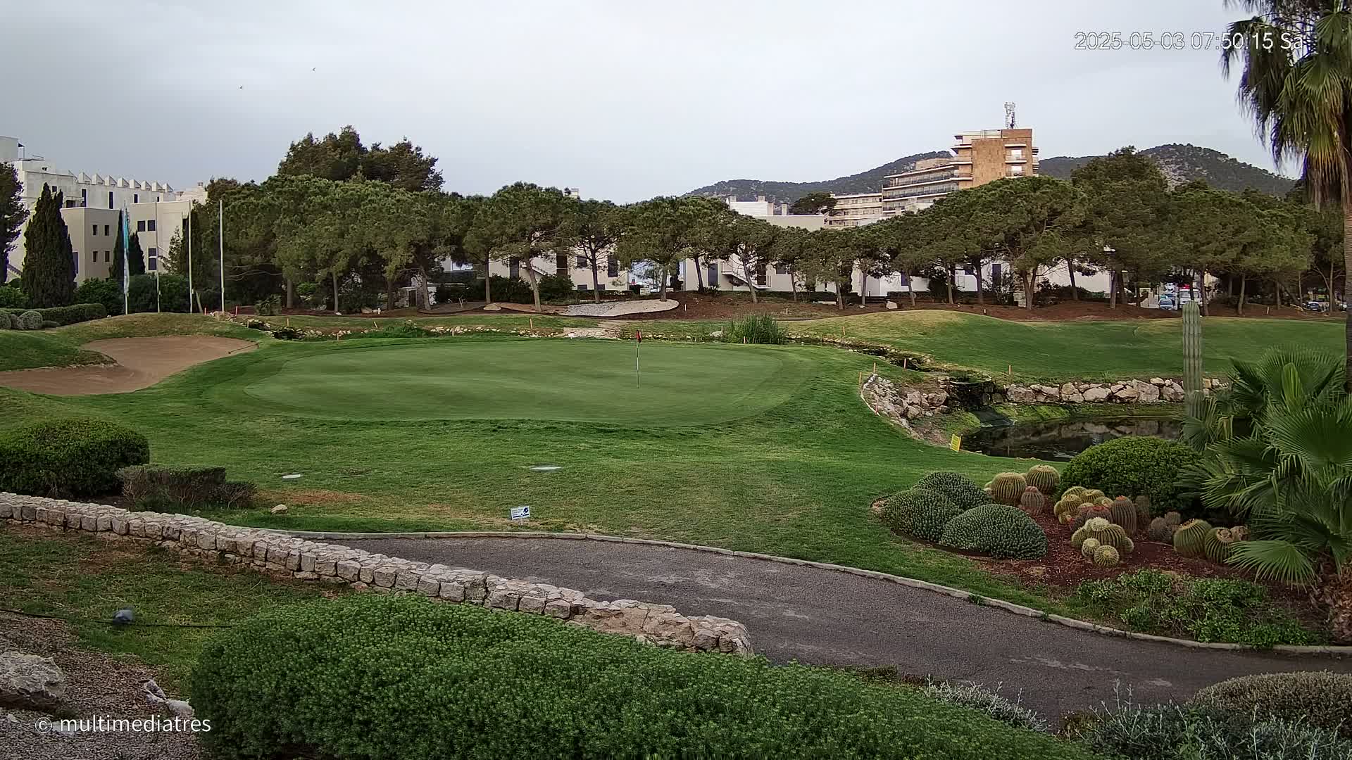 A golf course green with a sand trap, surrounded by trees, buildings, and a small pond under an overcast sky.