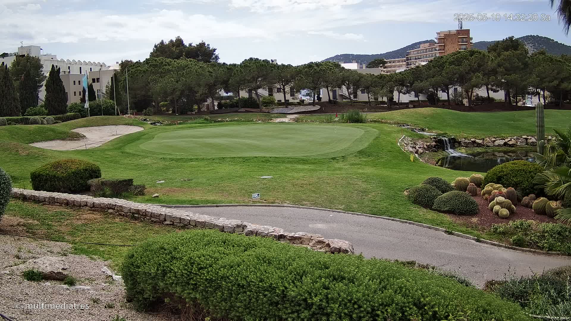 A golf course green with a sand trap, a small waterfall, and cacti, under a partly cloudy sky, is situated near buildings and trees.