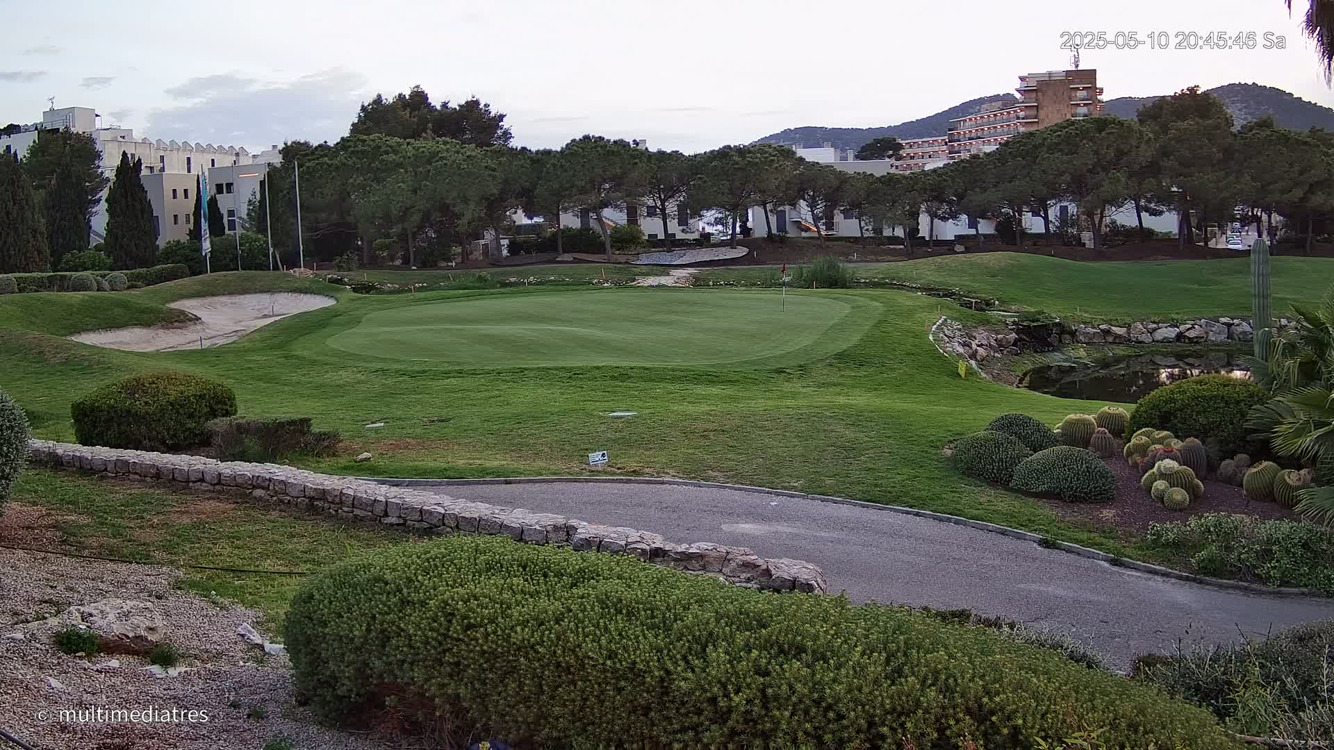 A golf course green with a sand trap and a small pond, bordered by shrubs, cacti, and buildings under a cloudy sky.