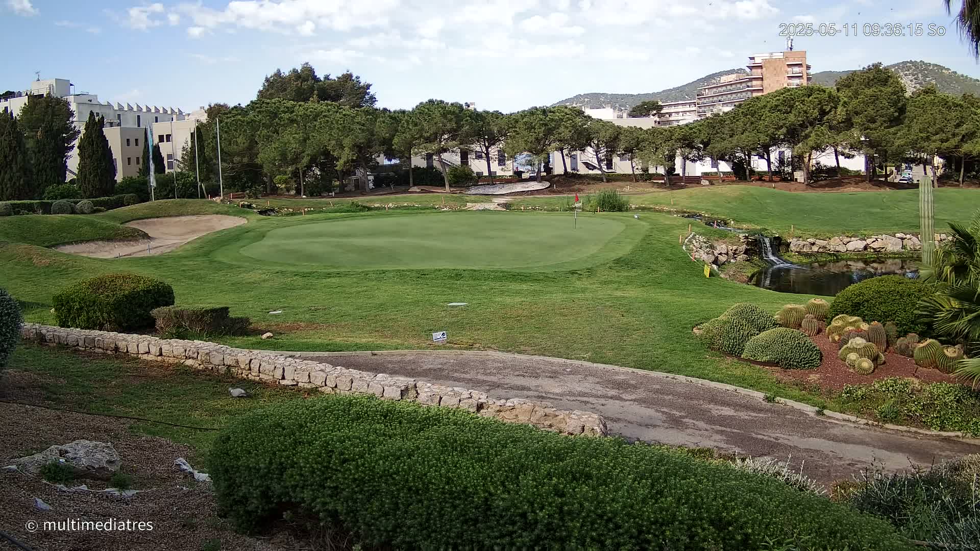 A golf course green with a sand trap, a small waterfall, and a collection of cacti is visible under a partly cloudy sky.