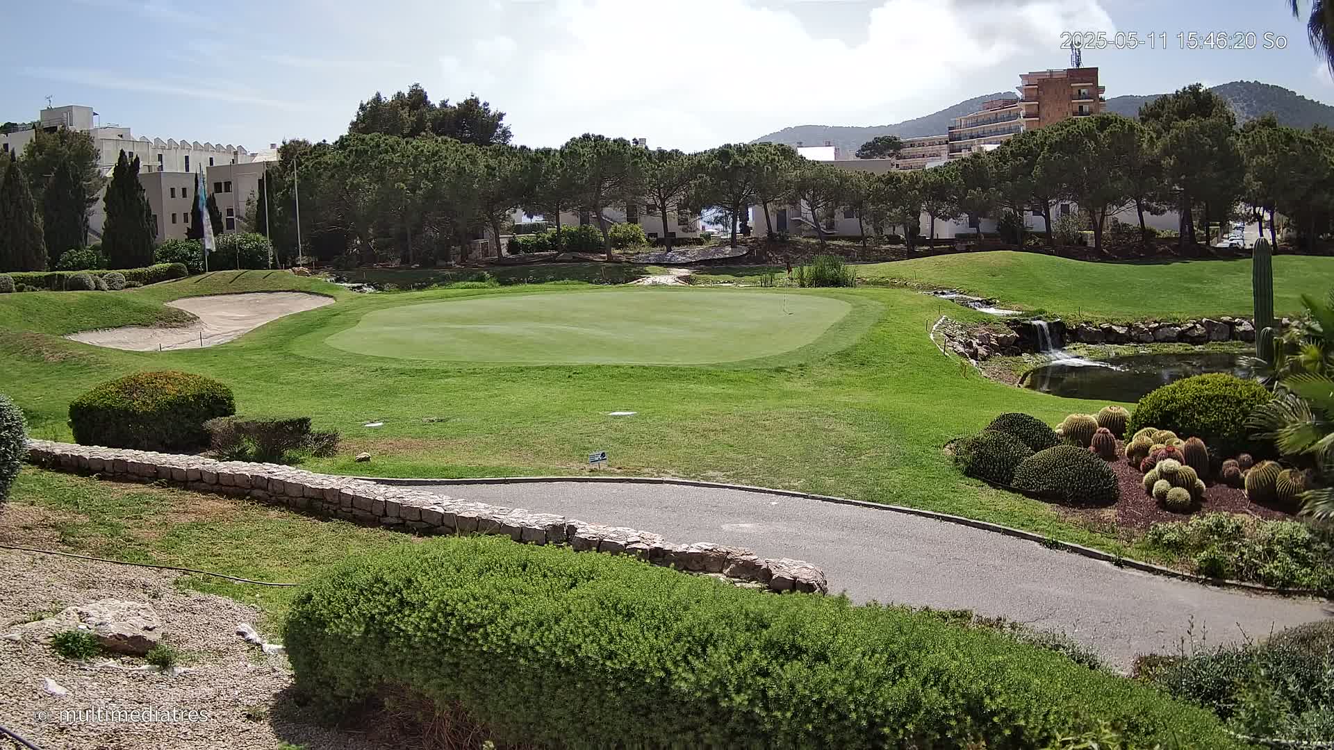 A golf course putting green with a sand trap, a small waterfall, and a variety of landscaping under a partly cloudy sky.