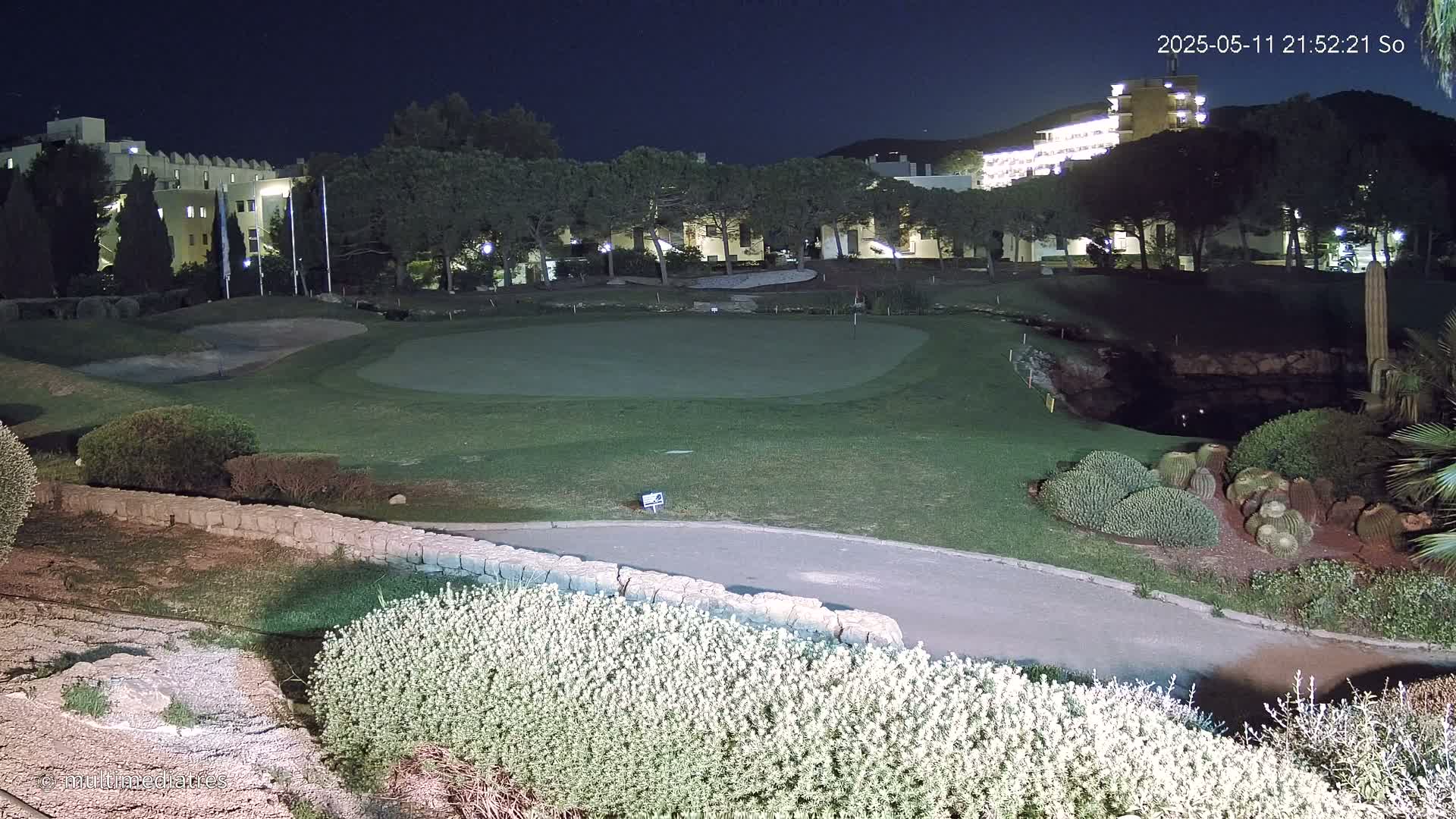 A putting green on a golf course is illuminated at night, with buildings and trees visible in the background.