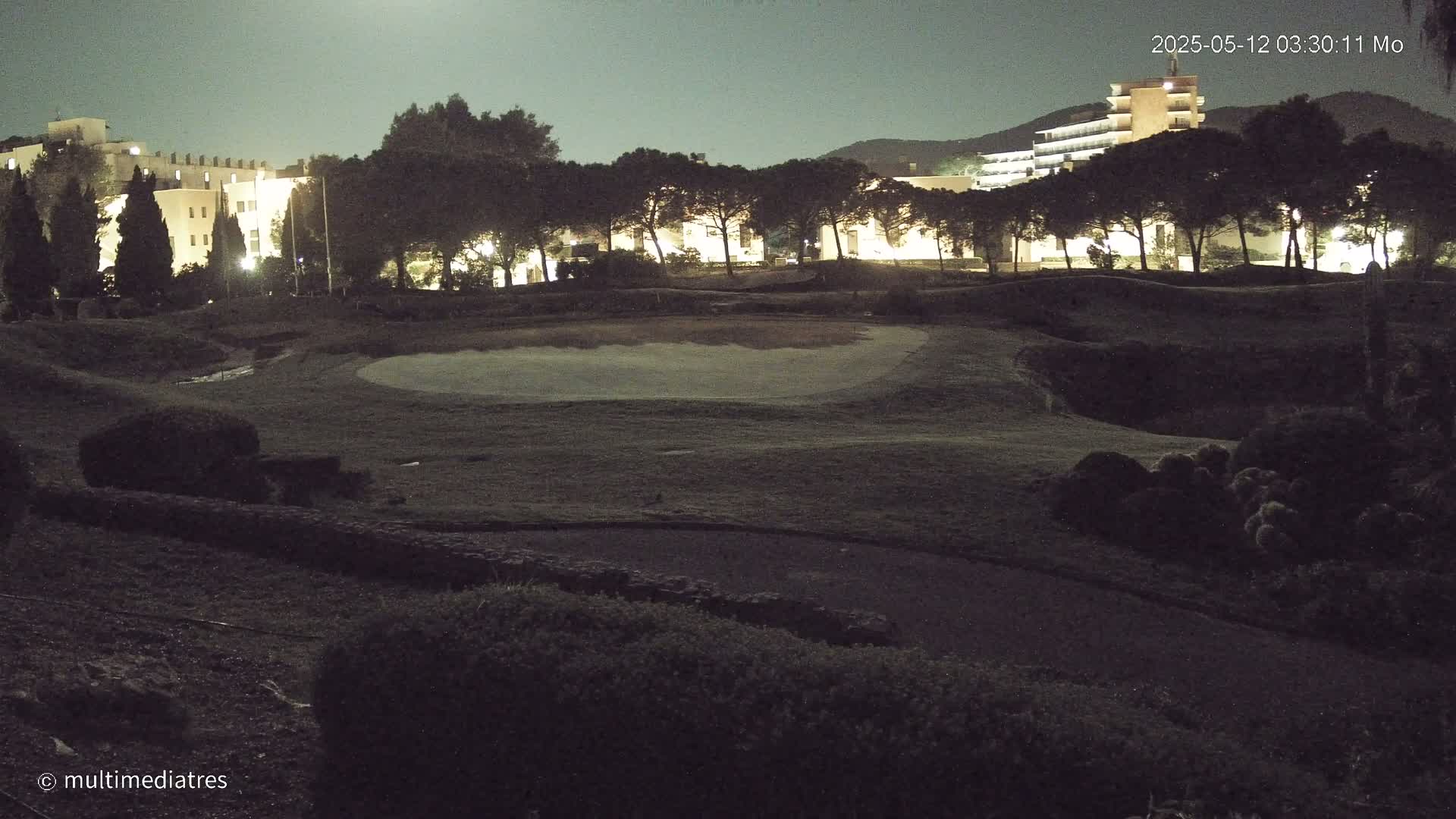 A night-time view of a golf course with buildings and trees illuminated under a dimly lit moon.