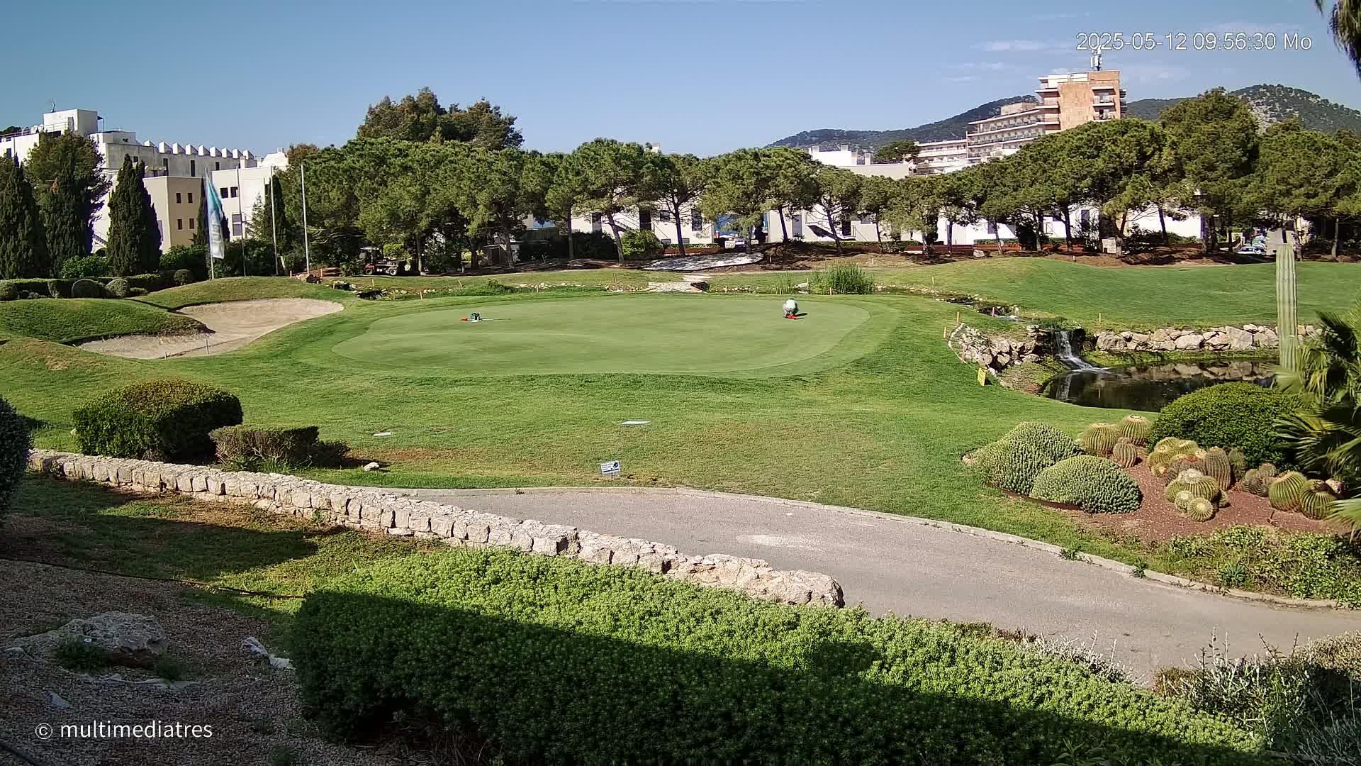 A golf course putting green with two golfers, a small pond, and buildings in the background under a clear sunny sky.
