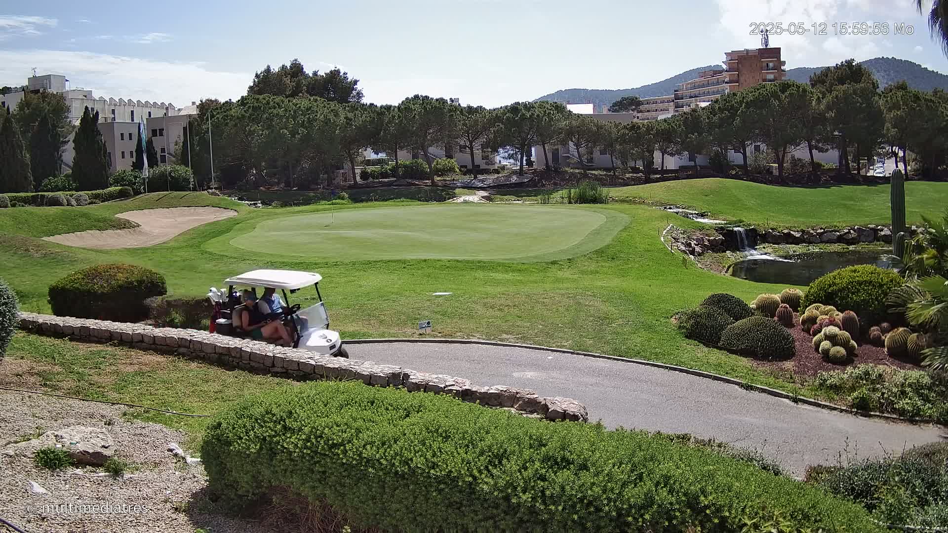 A golf cart with two people drives across a sunny golf course, past a small pond and manicured landscaping, with buildings visible in the background.