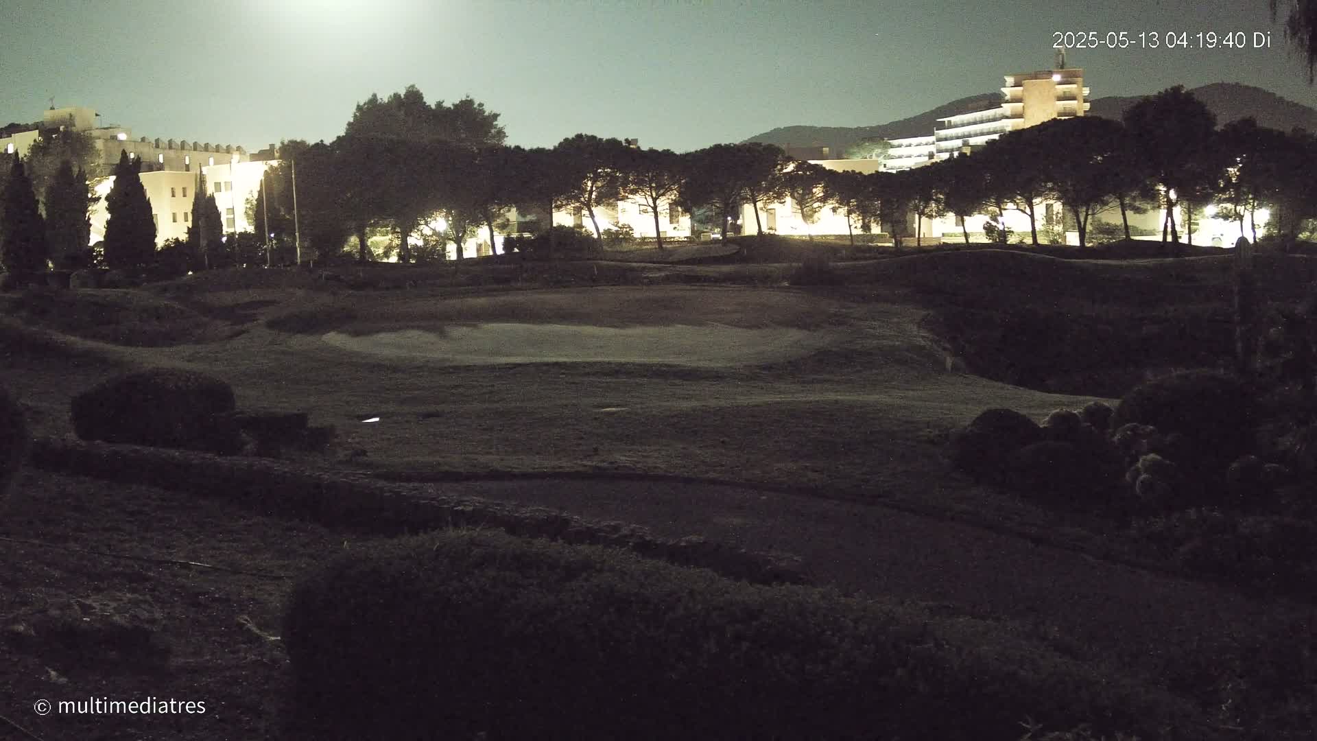 A nighttime view of a golf course with illuminated buildings in the background under a bright moon.