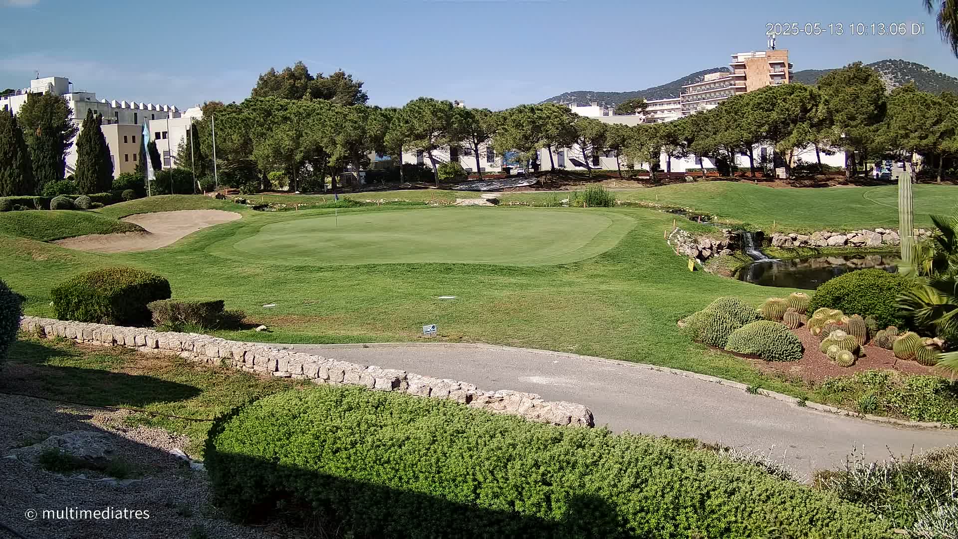 A golf course green, sand trap, and small pond are surrounded by manicured lawns, shrubs, and cactus plants under a clear, sunny sky, with buildings visible in the background.