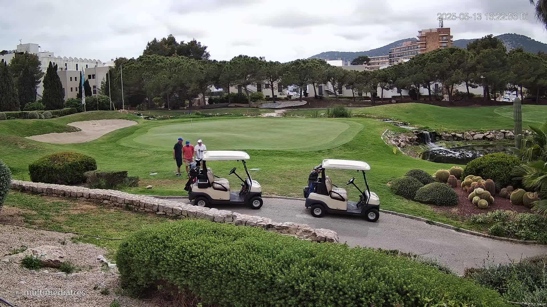 Two golf carts are parked near a putting green on a partly cloudy day, with three golfers nearby.