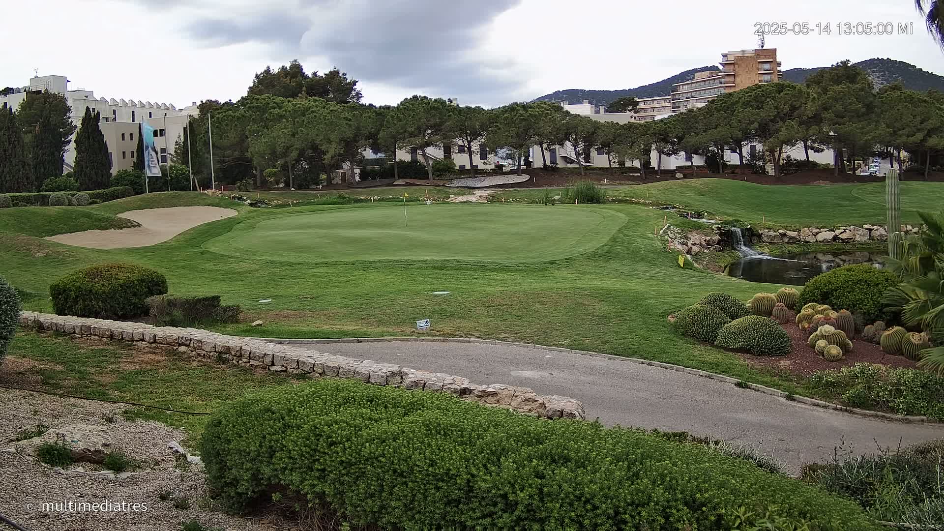 A golf course green with a sand trap, a small waterfall, and several cacti, under a cloudy sky.