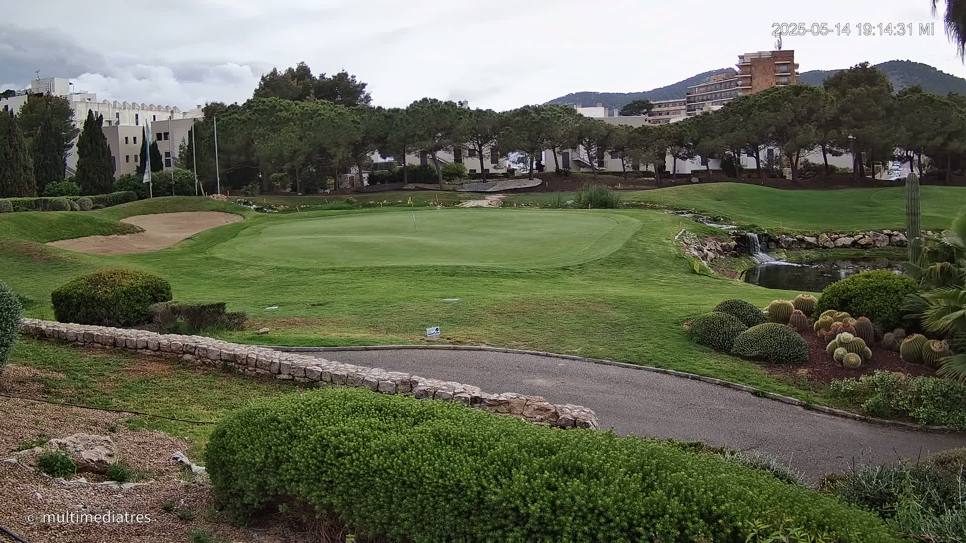 A golf course green with a sand trap, a small waterfall, and a pond, under a cloudy sky, is situated near buildings and trees.