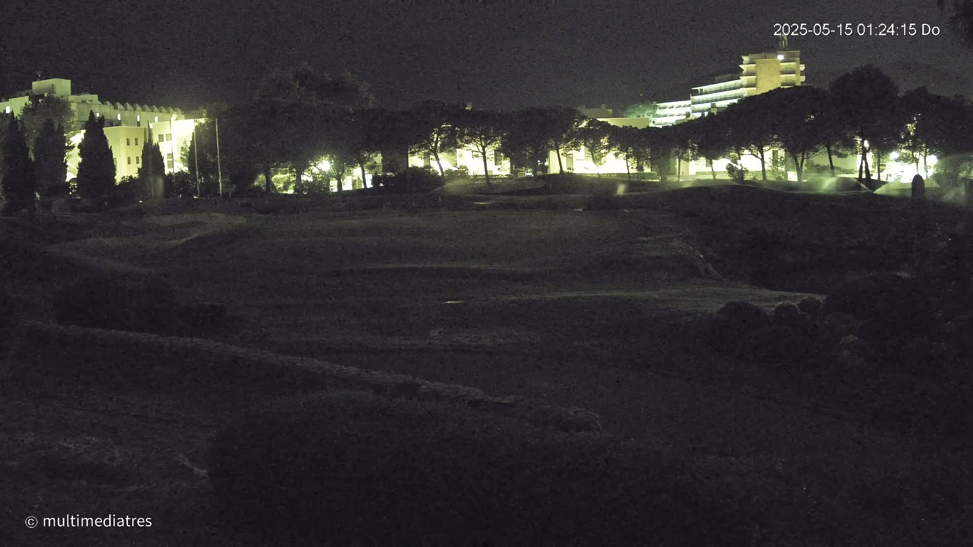A dark, nighttime view of a grassy area with buildings and trees illuminated in the background.