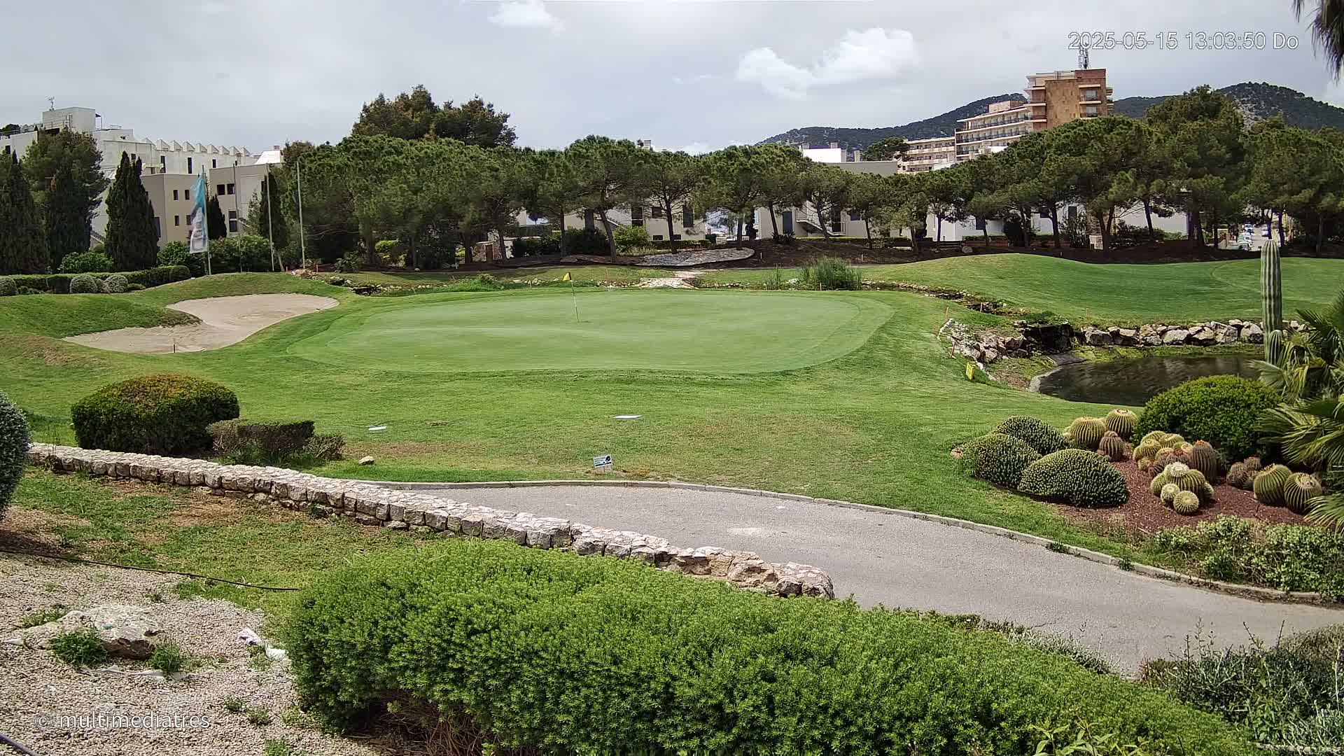 A golf course green, sand trap, and landscaping featuring a small pond and cacti are visible under a partly cloudy sky.