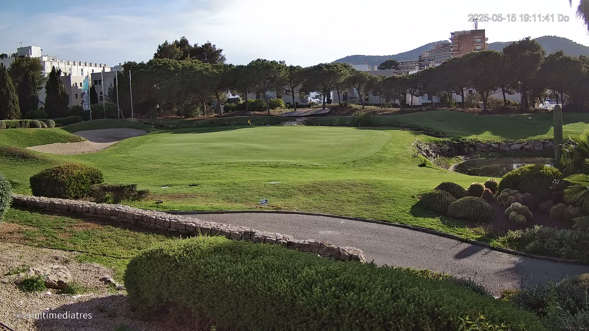 A golf course green with sand traps, a paved path, and low shrubs, under sunny skies, is situated near buildings and trees.