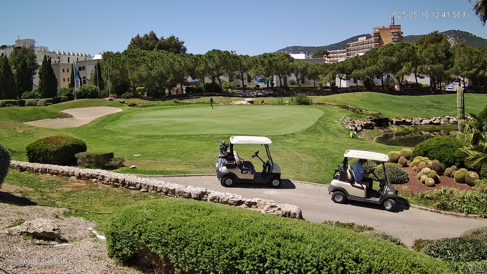 Two golf carts are parked on a path next to a putting green on a sunny day, with a person visible in the distance on the green.