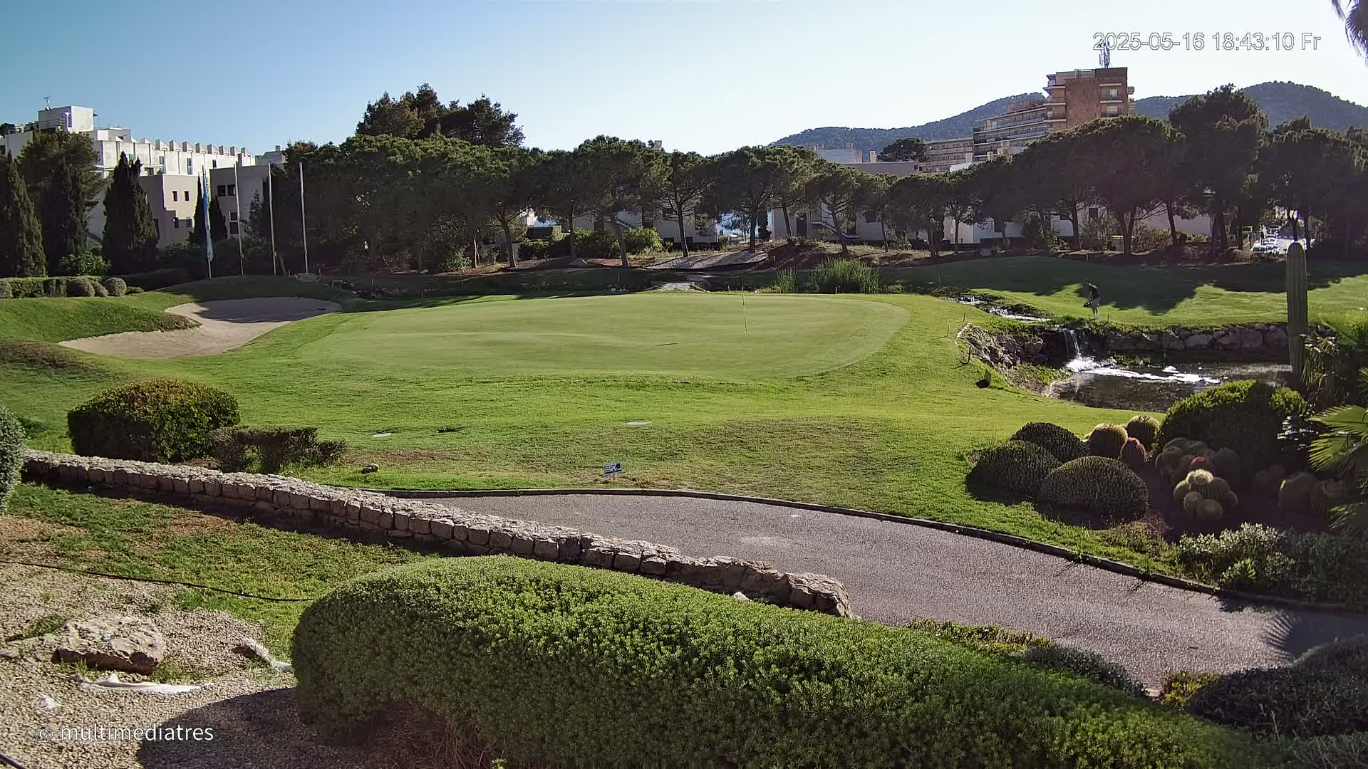 A golf course green with a sand trap, a small waterfall, and a paved path, all under a clear, sunny sky.