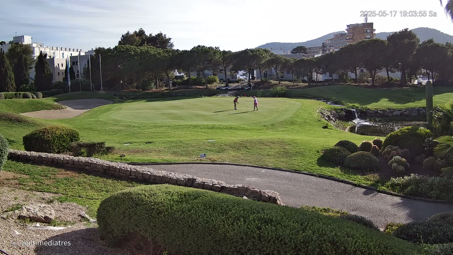 Two golfers are on a putting green surrounded by landscaping, a small waterfall, and buildings under a partly sunny sky.