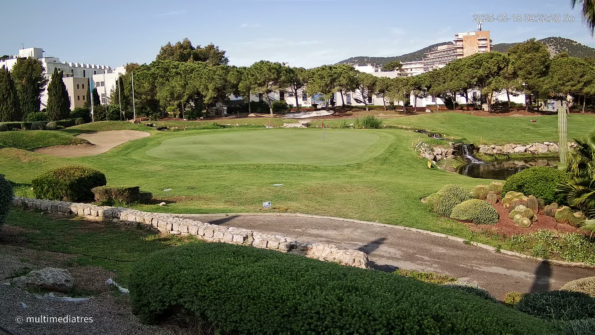 A golf course green, sand trap, and small pond are situated near buildings and a hillside under a clear sky.