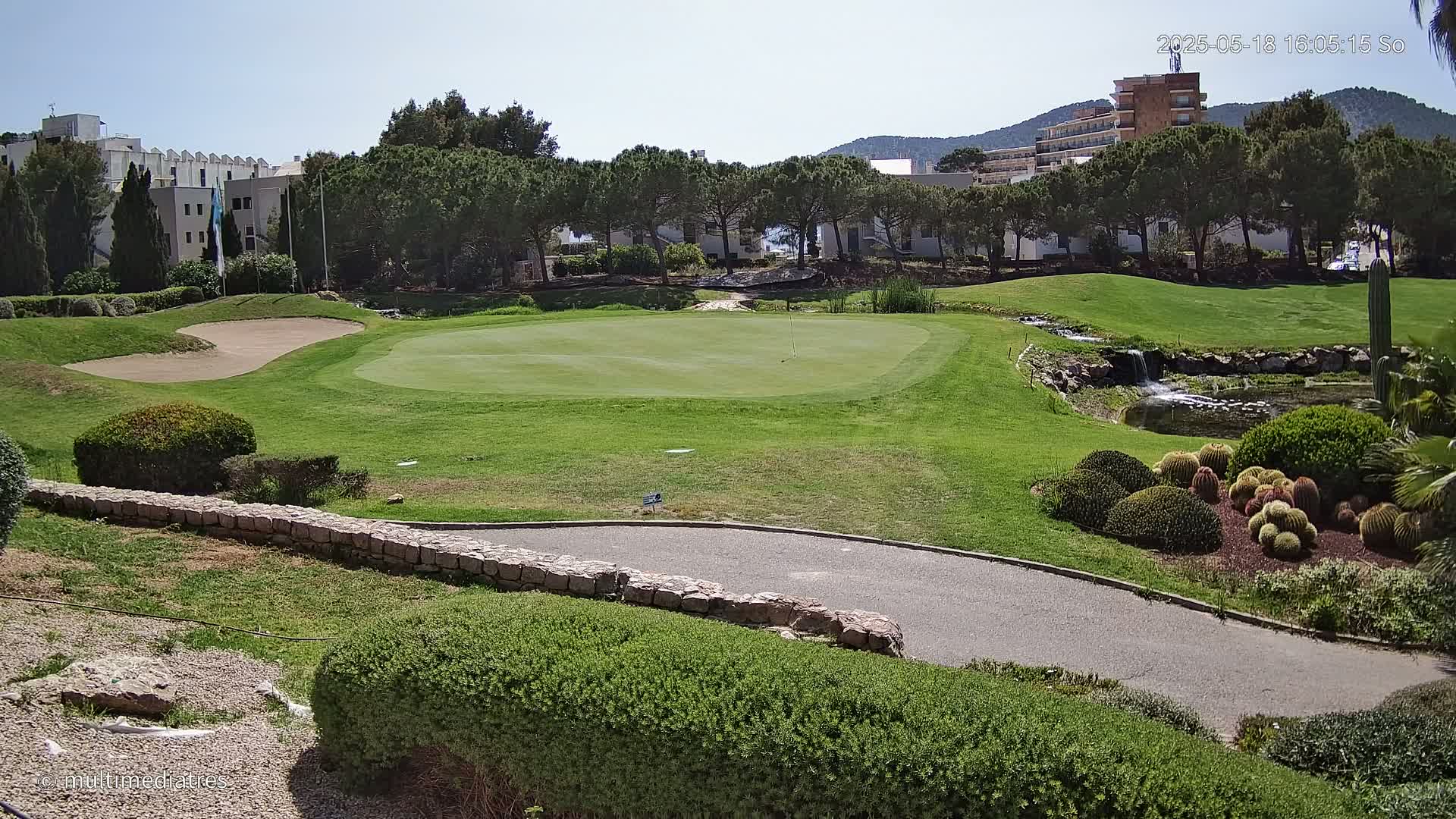 A sunny view of a golf course green, sand trap, and a paved path, with a small waterfall and various shrubs and cacti surrounding it, all set against a backdrop of buildings and trees under a clear sky.